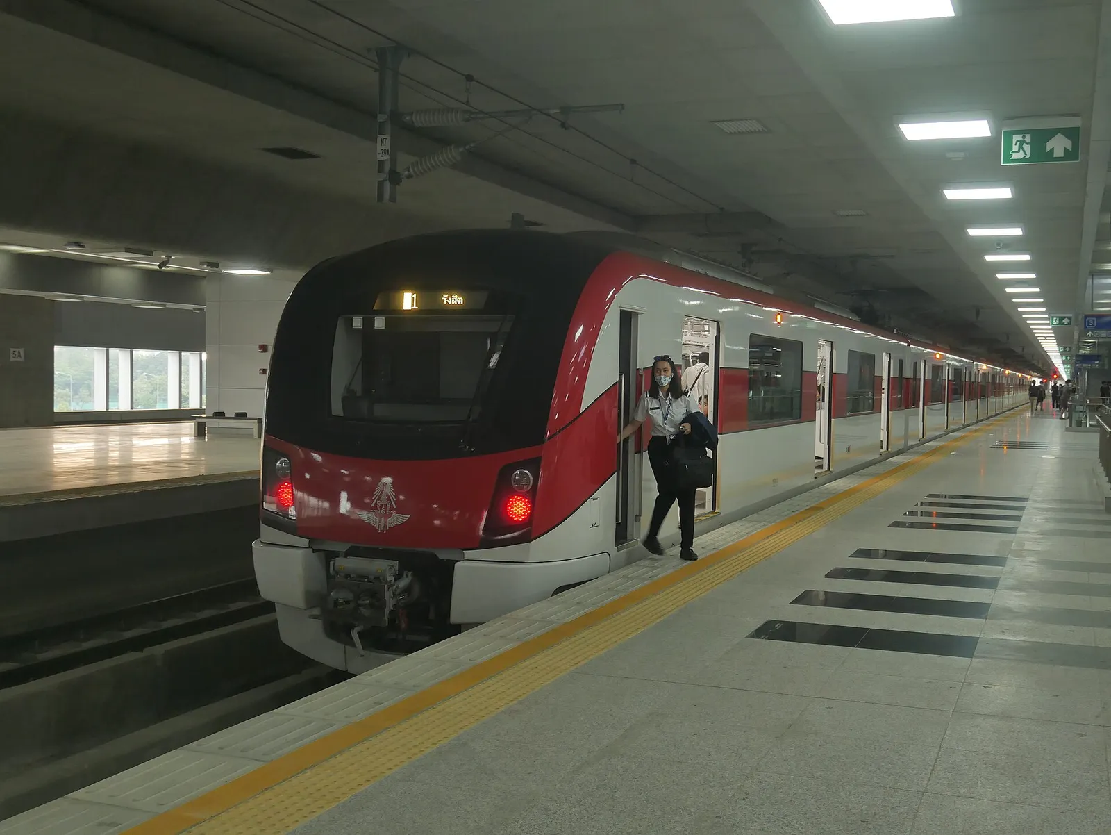 Red and white electric train beside a platform with yellow tactile paving, and a staff member at an open door.