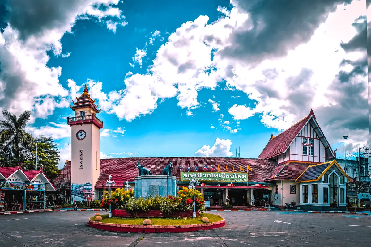 Red-roofed station building with a clock tower behind a flower bed and a traffic circle under a blue sky.