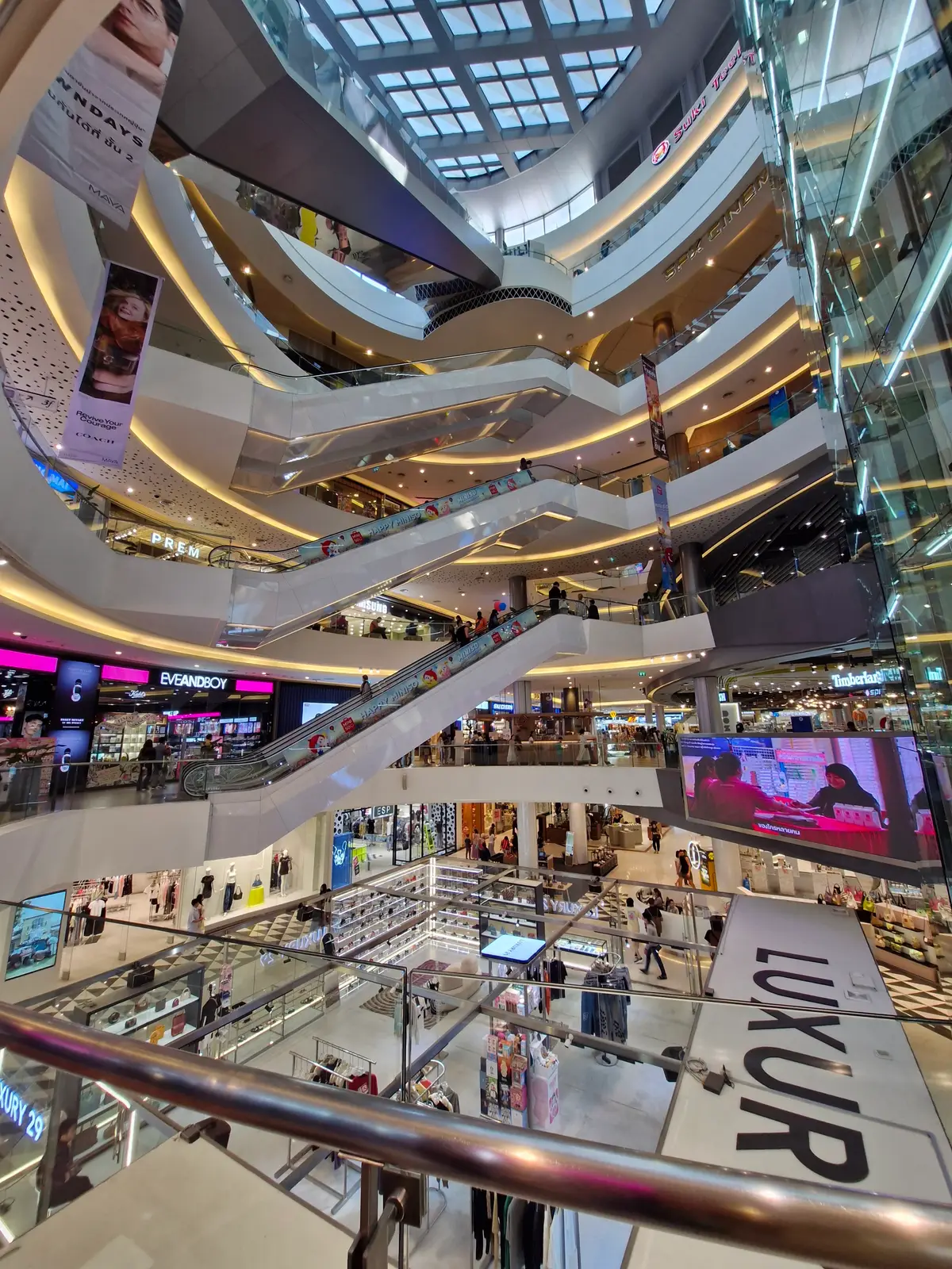 Escalators crossing a bright mall atrium with shop signs and a large video screen on the lower level.