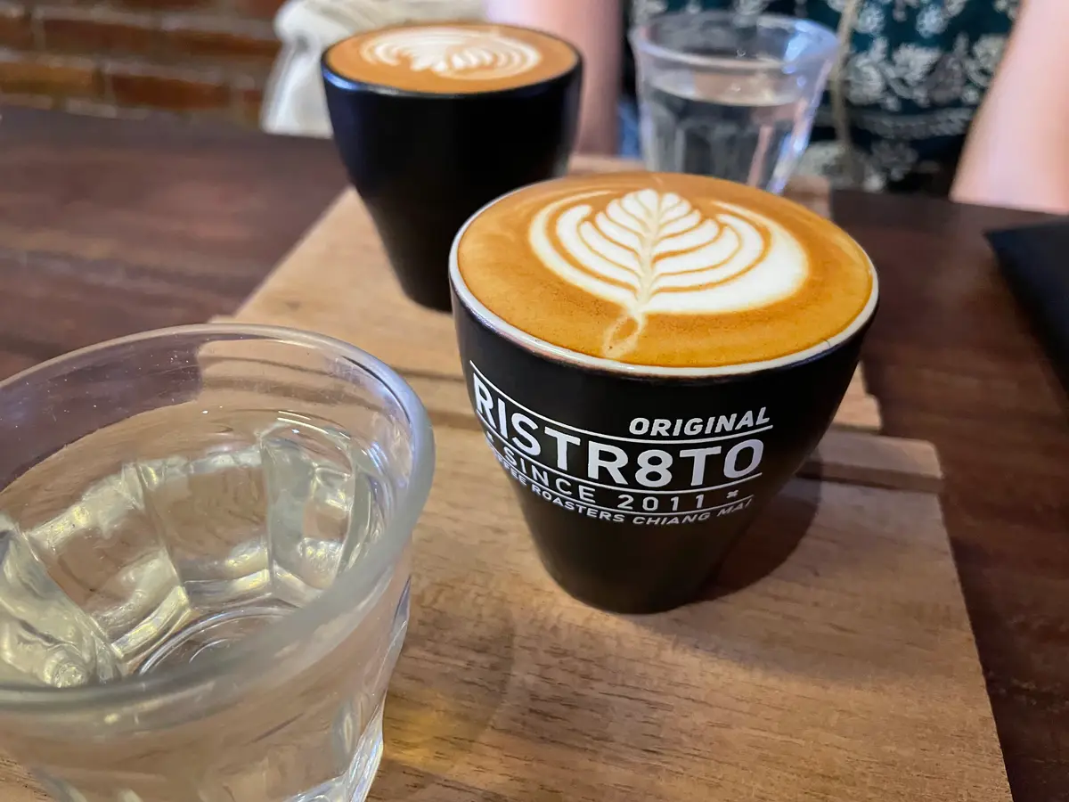 A black Ristr8to cup with leaf latte art on a wooden board, next to a glass of water.