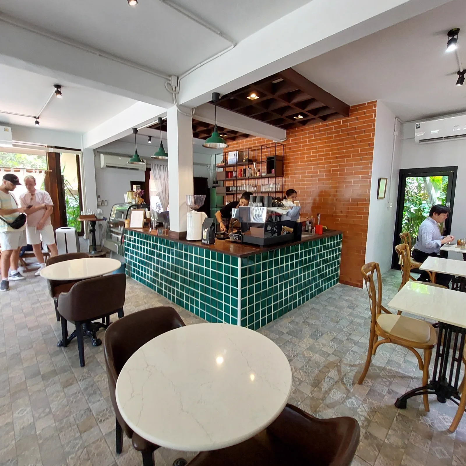 Interior with a green-tiled counter, brick wall and pendant lamps, baristas far back and a few guests seated