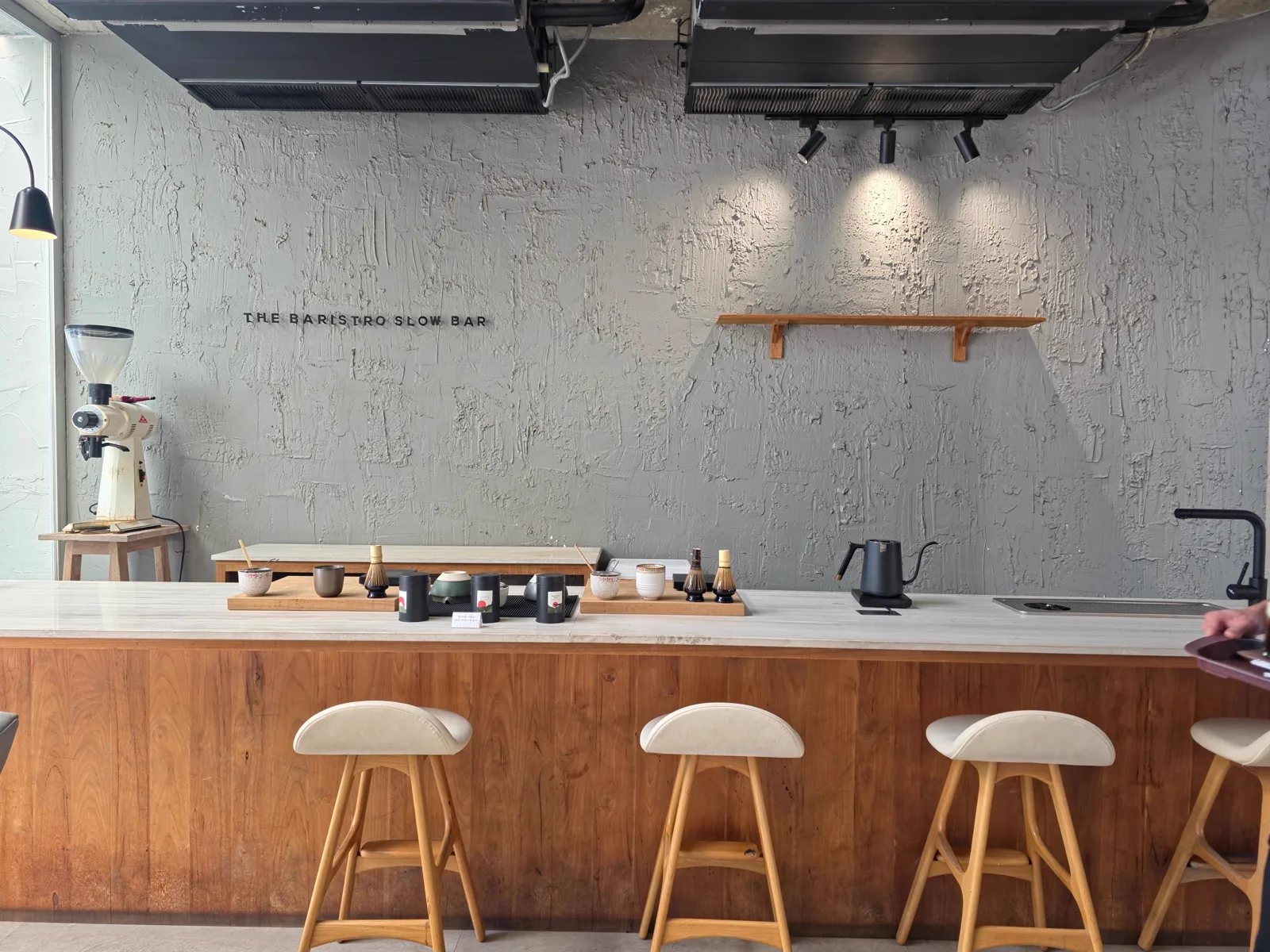 Matcha bar with a wooden front, cream bar stools, matcha tins and wall text The Baristro Slow Bar
