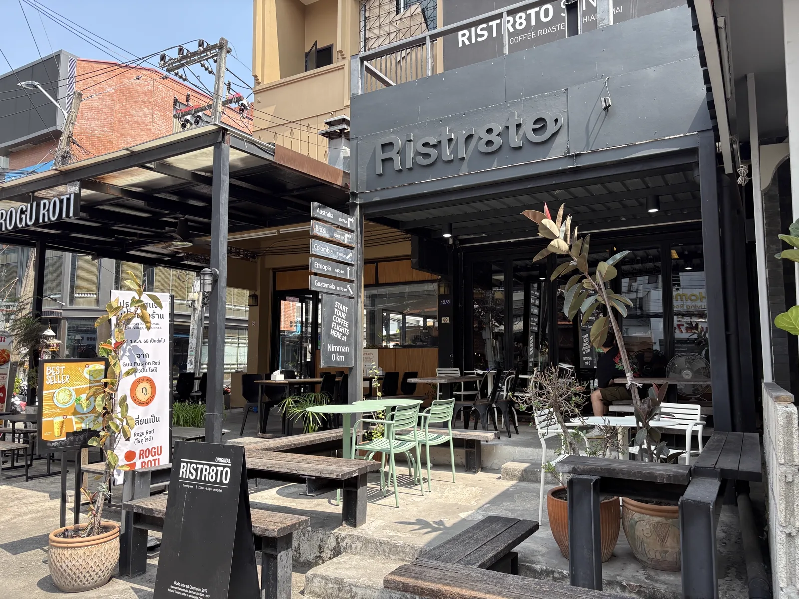 Dark storefront with a Ristr8to sign, direction boards and green chairs on the terrace