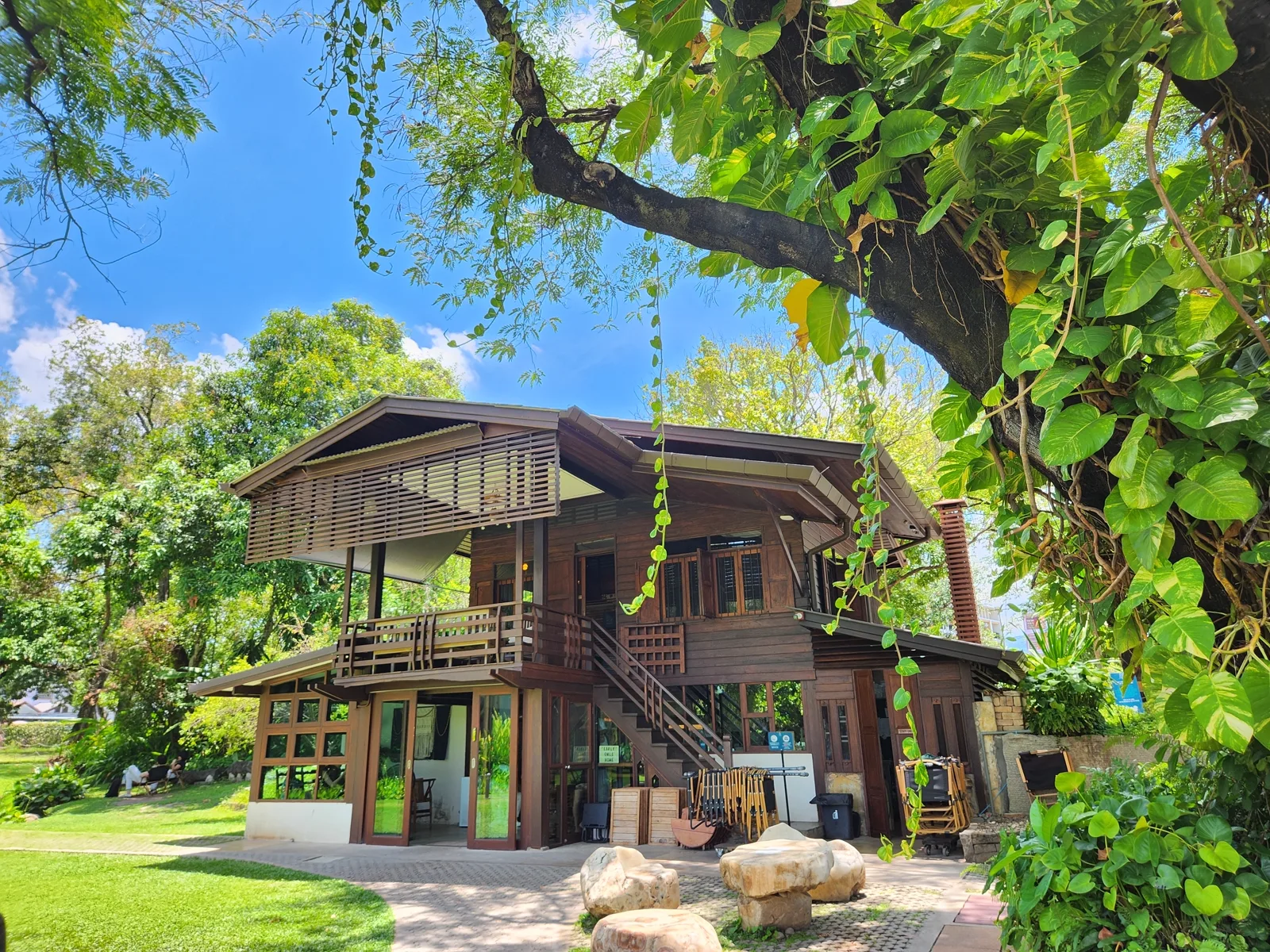 Dark wooden two-storey house with a slatted awning, balcony, large tree and carved stone stools in front