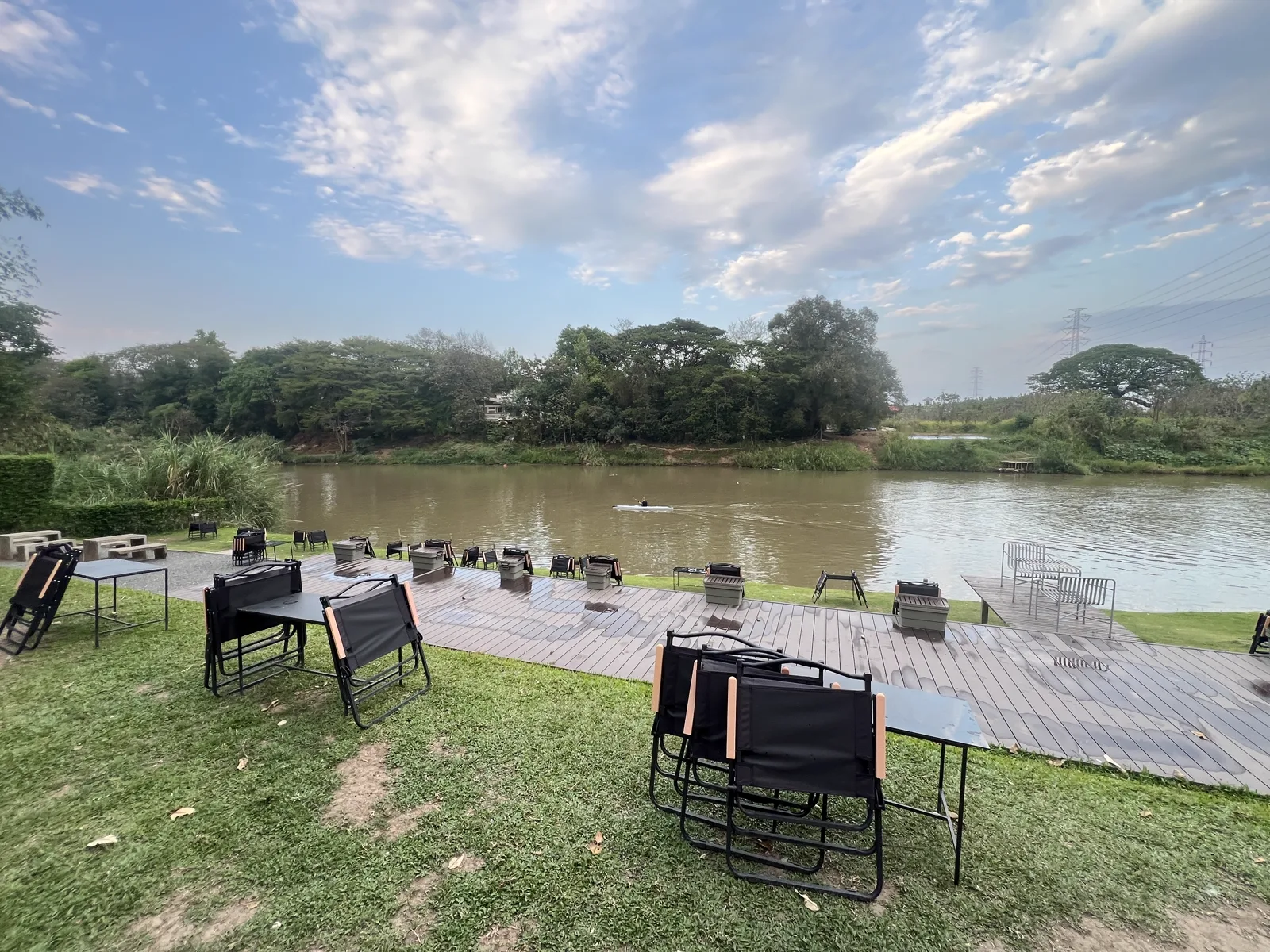 Riverside wooden deck with folding chairs and tables above a brown river, a distant kayak and trees opposite
