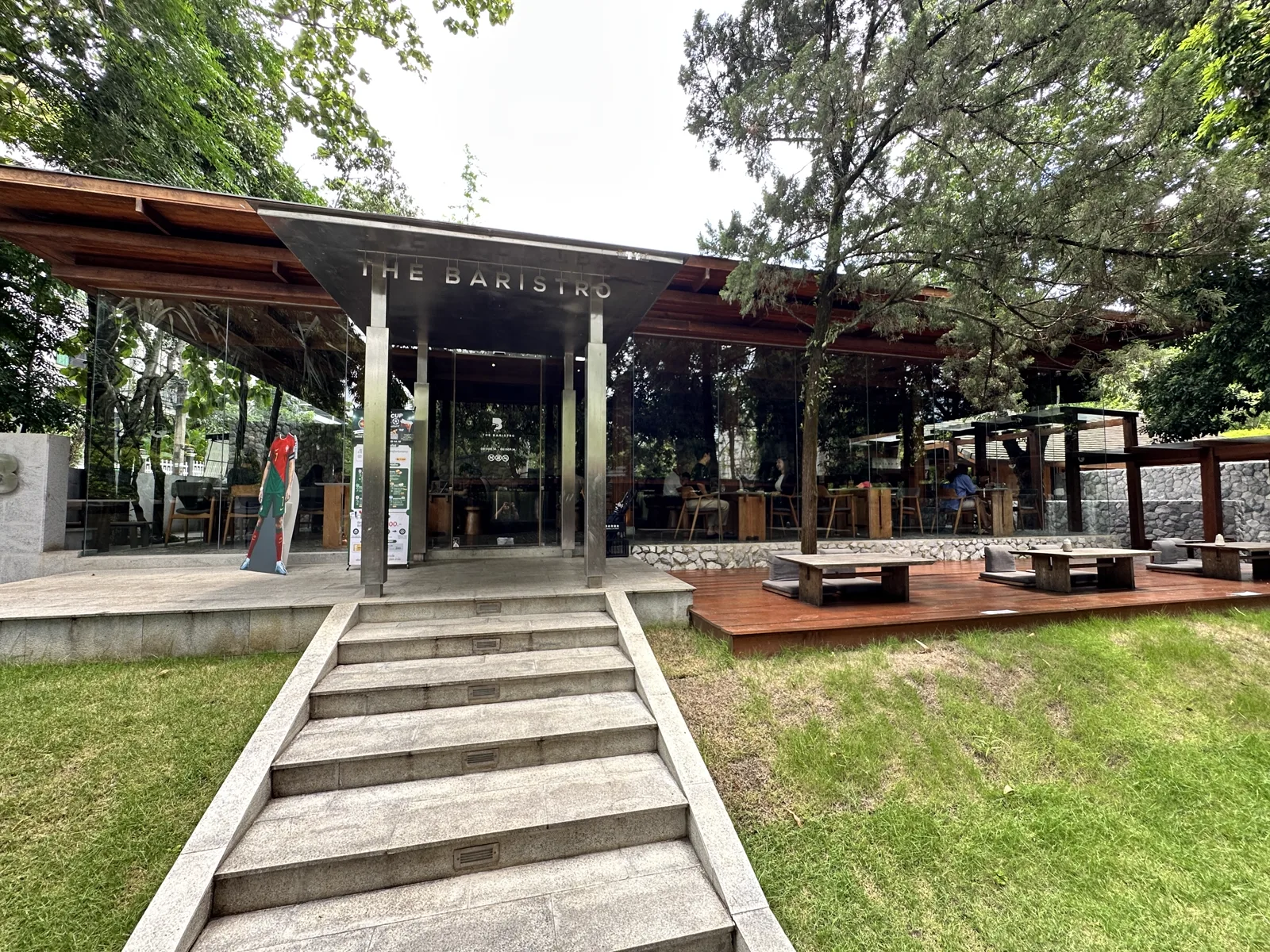 Cafe front under a metal canopy with Baristro lettering, stone steps, glass walls and wooden tables