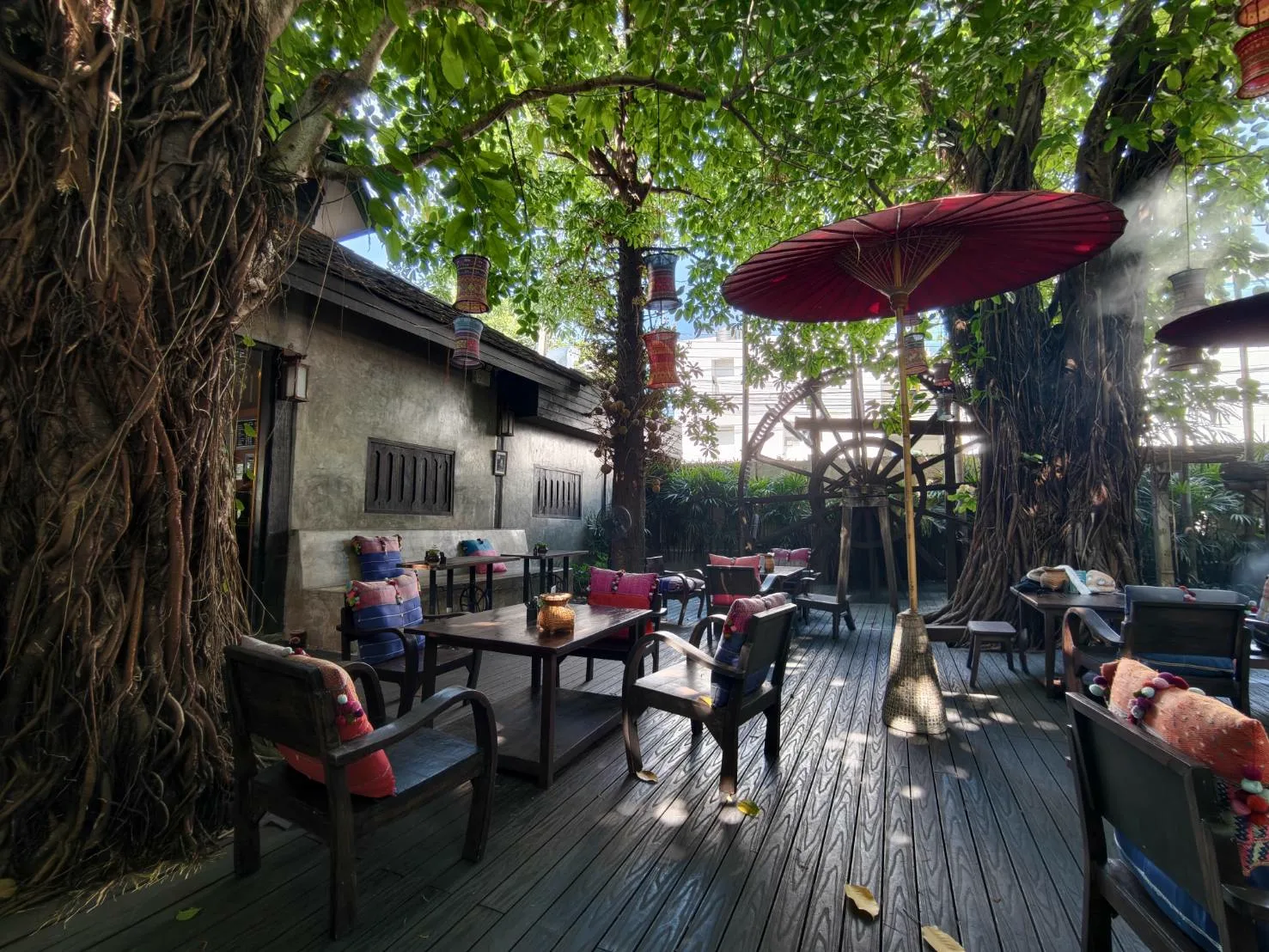 Outdoor deck under large banyan trees with a red umbrella, lanterns and a water wheel