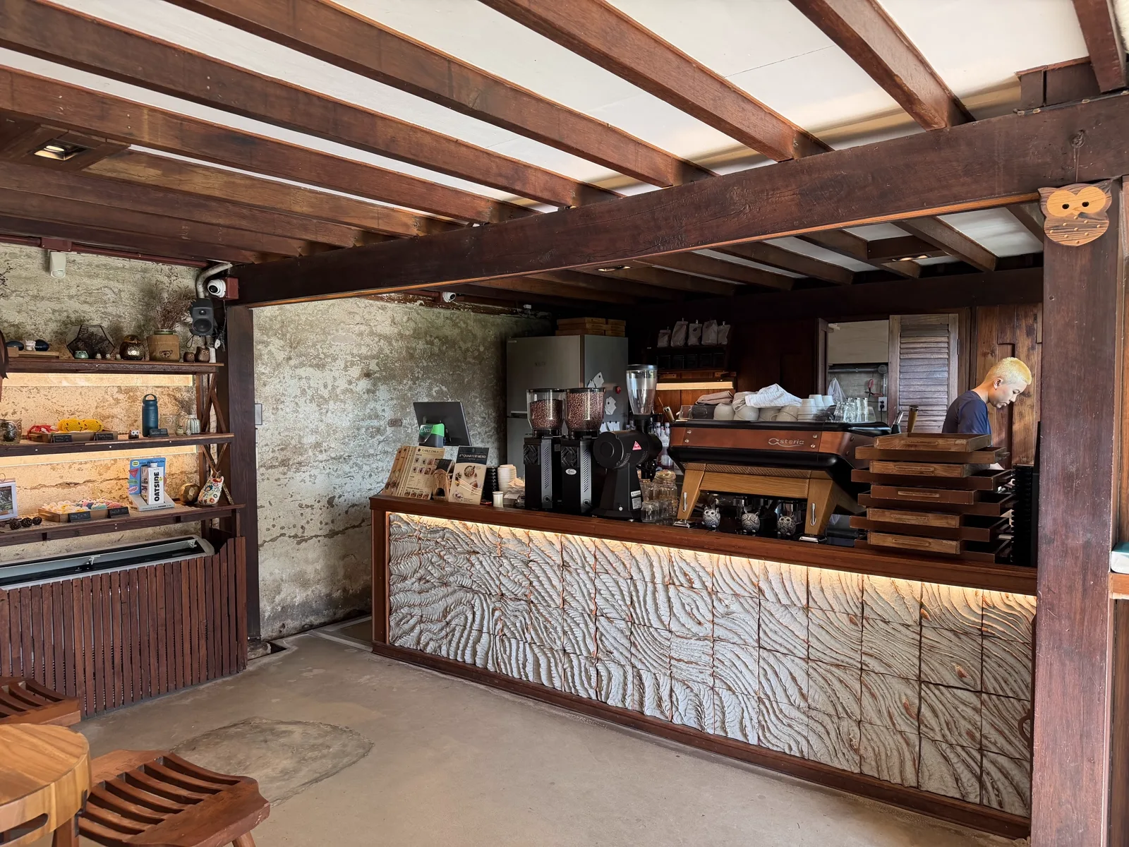 Indoor coffee bar with an owl-carved front panel, espresso machine, shelves of goods and one barista