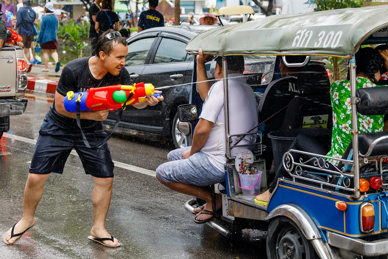 Man aiming a red and blue water gun on a wet street beside a three-wheeled tuk-tuk.