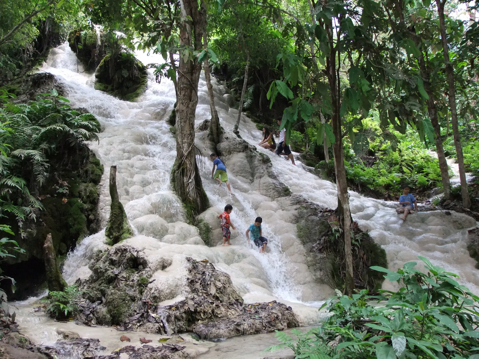 White limestone cascade running down through forest with a few climbers on the rock