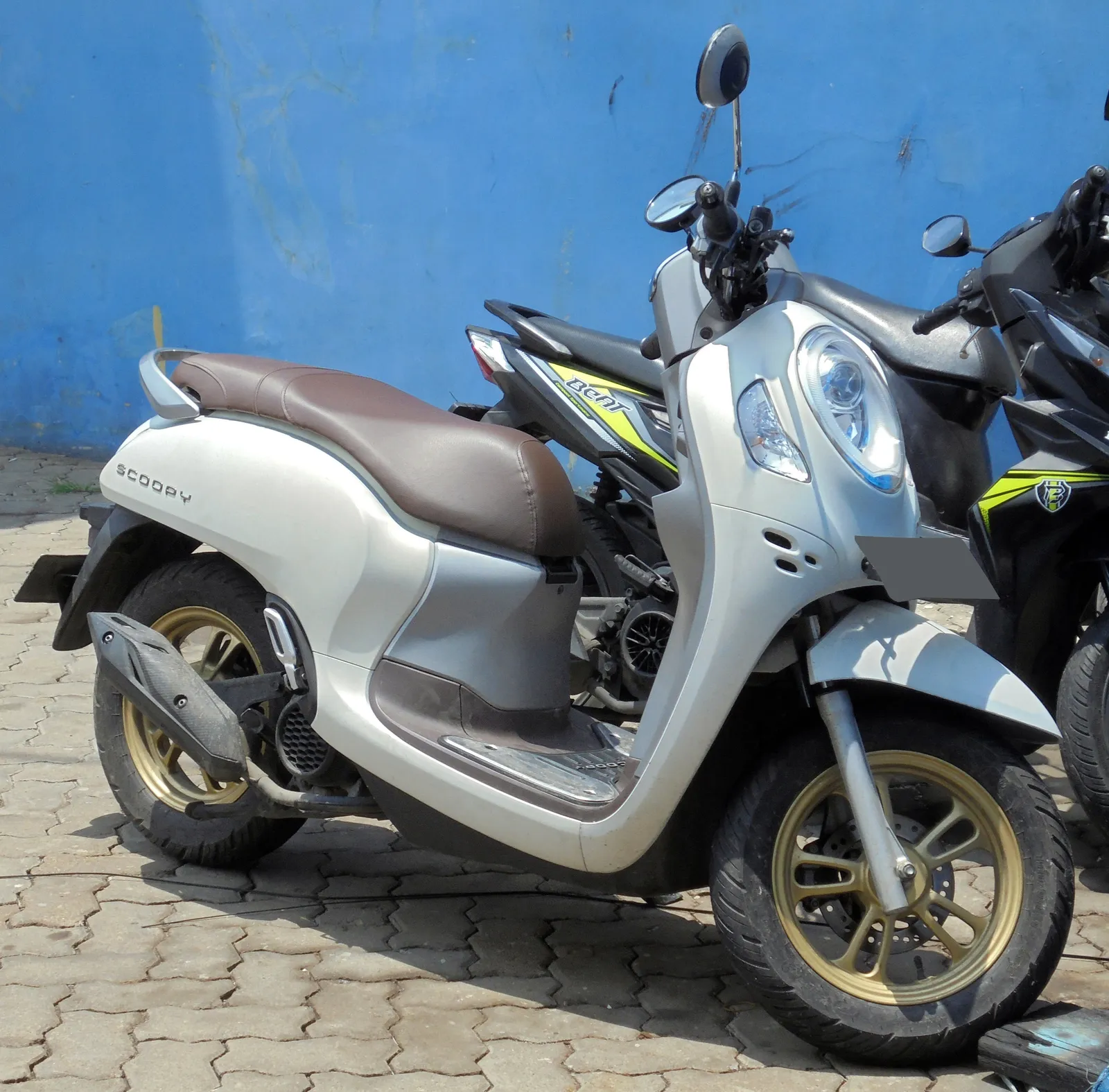 Silver and brown Honda Scoopy scooter parked on paving beside a blue wall.