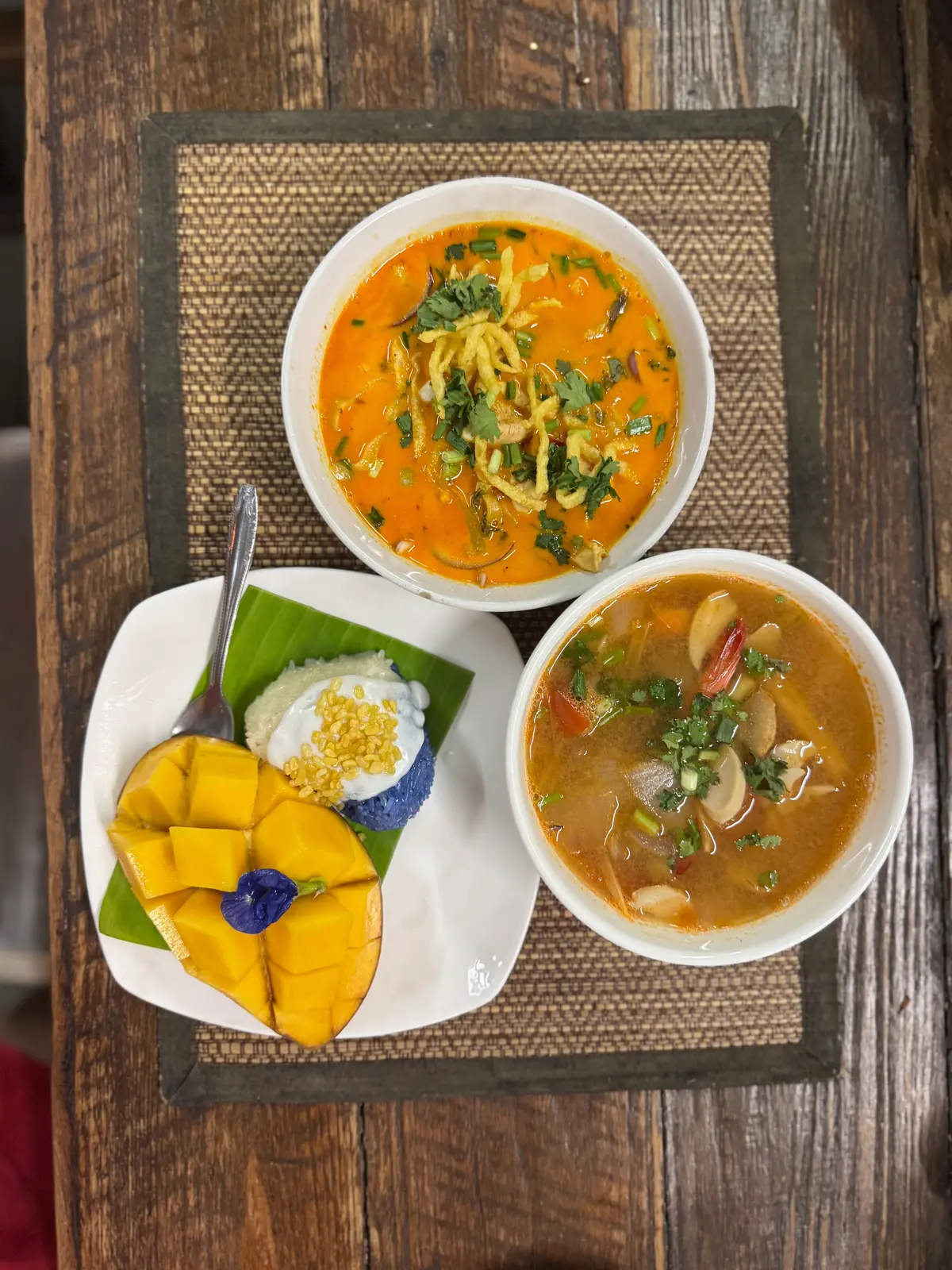 Bowls of orange curry noodle soup and hot and sour soup beside mango with sticky rice on a banana leaf.