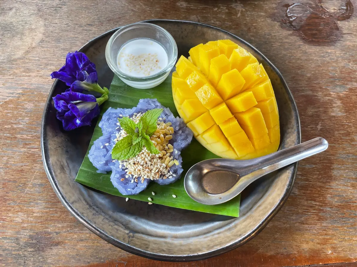 Mango and blue sticky rice on a banana leaf on a dark plate with a spoon.