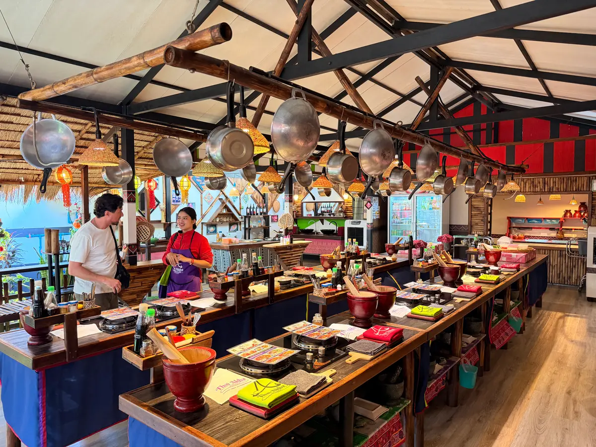 A cooking counter lined with woks and mortars under hanging pans, with two people at the far end.