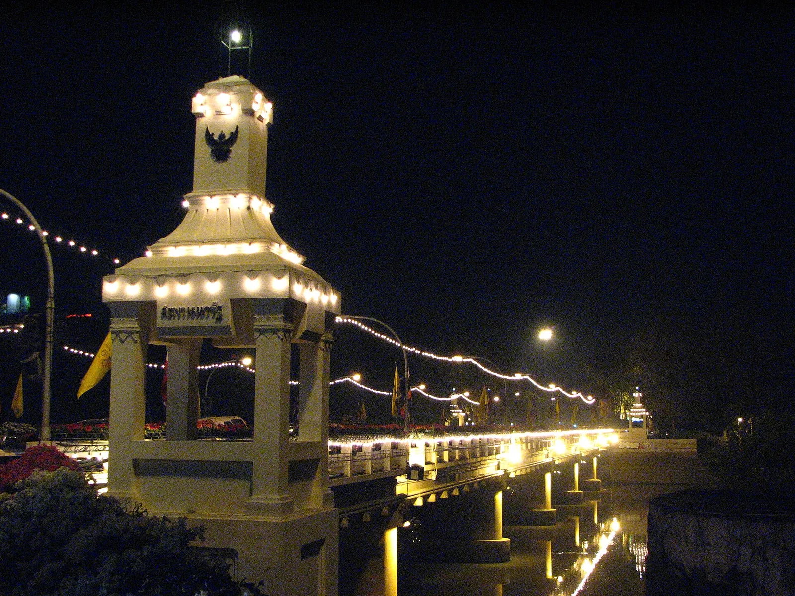 Night view of a bridge strung with lights beside a lit gate tower topped with a bird emblem.