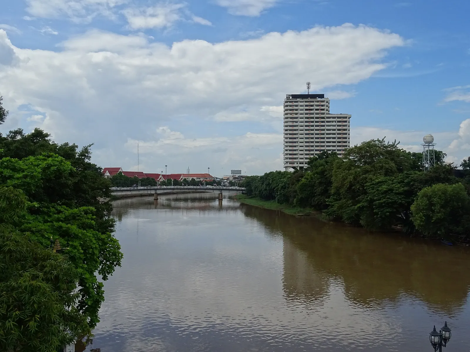 Brown river flowing between green banks with a tall hotel block and a bridge in the distance under a cloudy sky.
