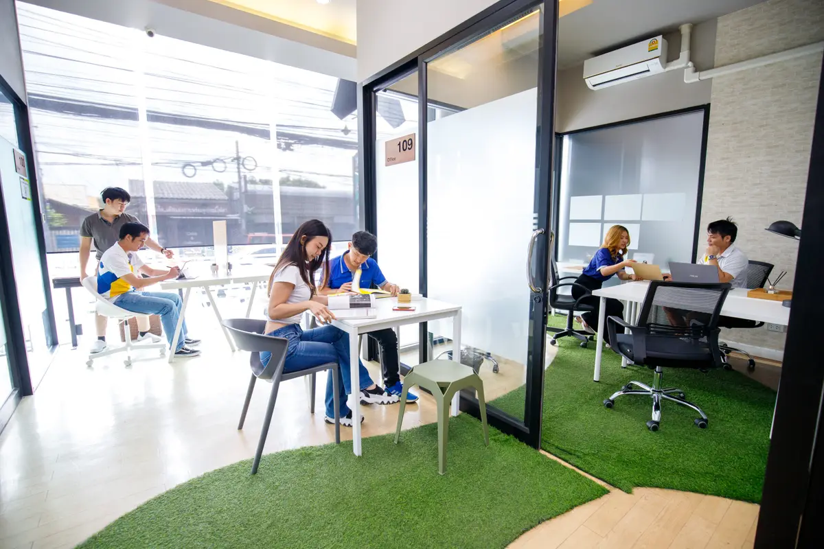 Four people working at a white table in a glass-walled room with green artificial turf on the floor.