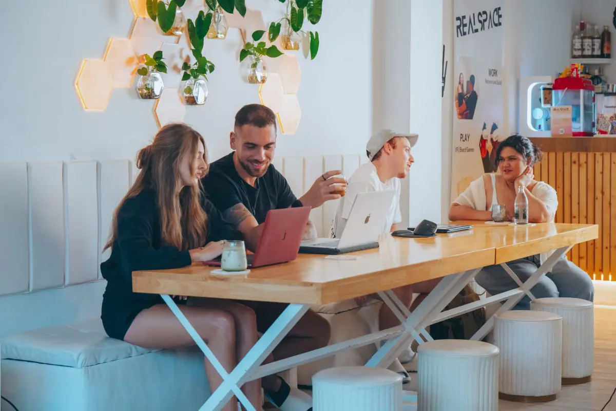 Four people talking over laptops at a long wooden table in a white room.
