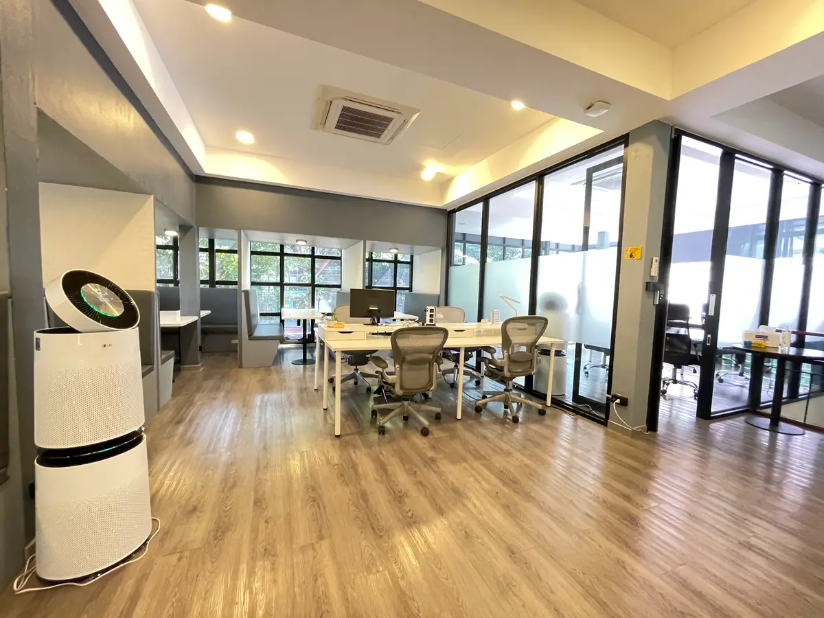 Wide room with a wooden floor, long meeting tables, office chairs and an air purifier beside tall windows.