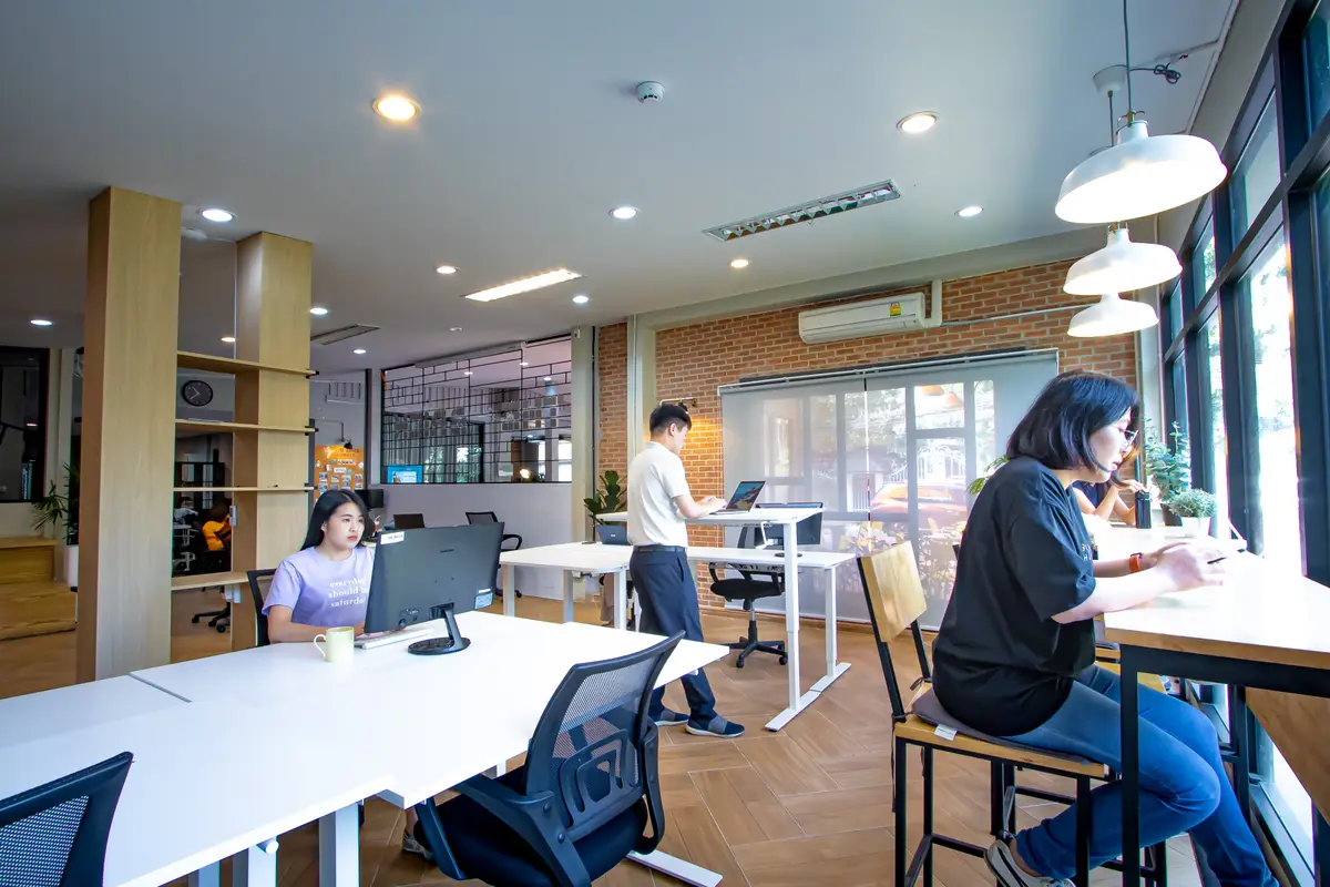 People working at white desks under pendant lamps in a room with a brick wall.
