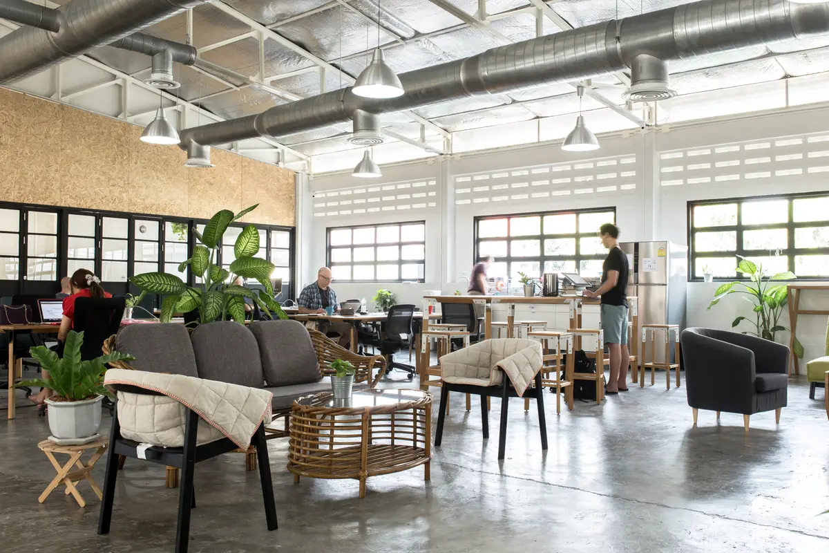 Open industrial room with sofas, plants, desks and hanging lamps where people work.
