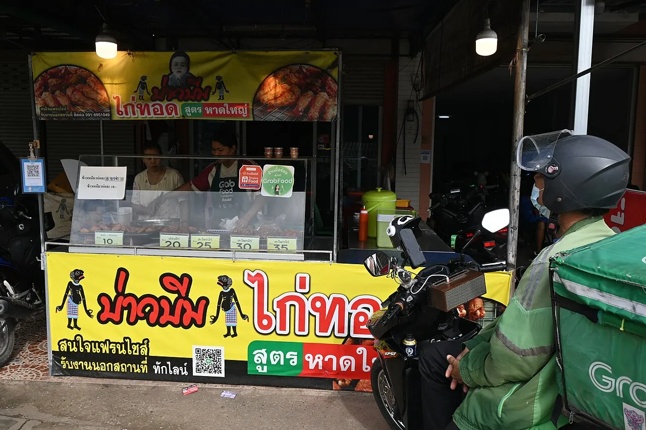 A delivery rider in a green jacket on a motorbike beside a yellow street food stall with a Thai sign