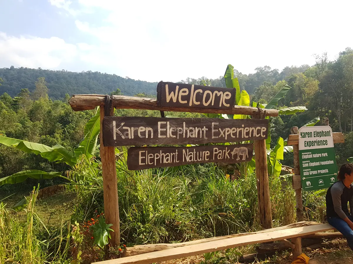 A wooden welcome sign reading Karen Elephant Experience, Elephant Nature Park, beside a path.
