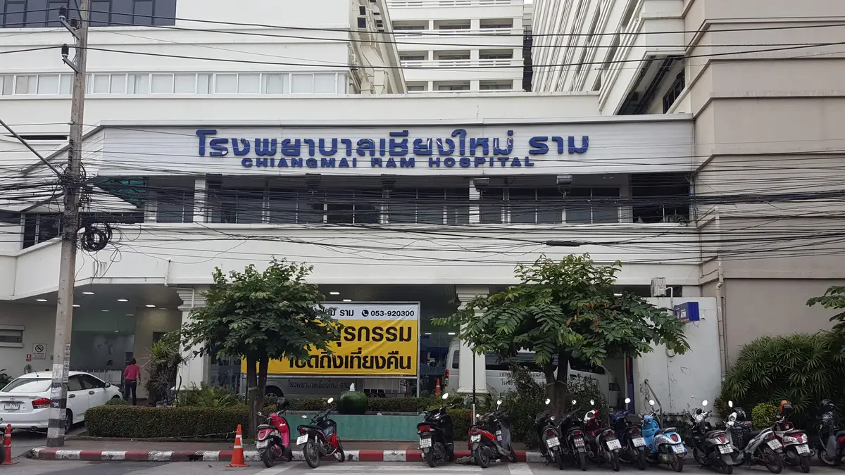 Hospital front with a blue Thai and English name sign above a row of parked motorbikes