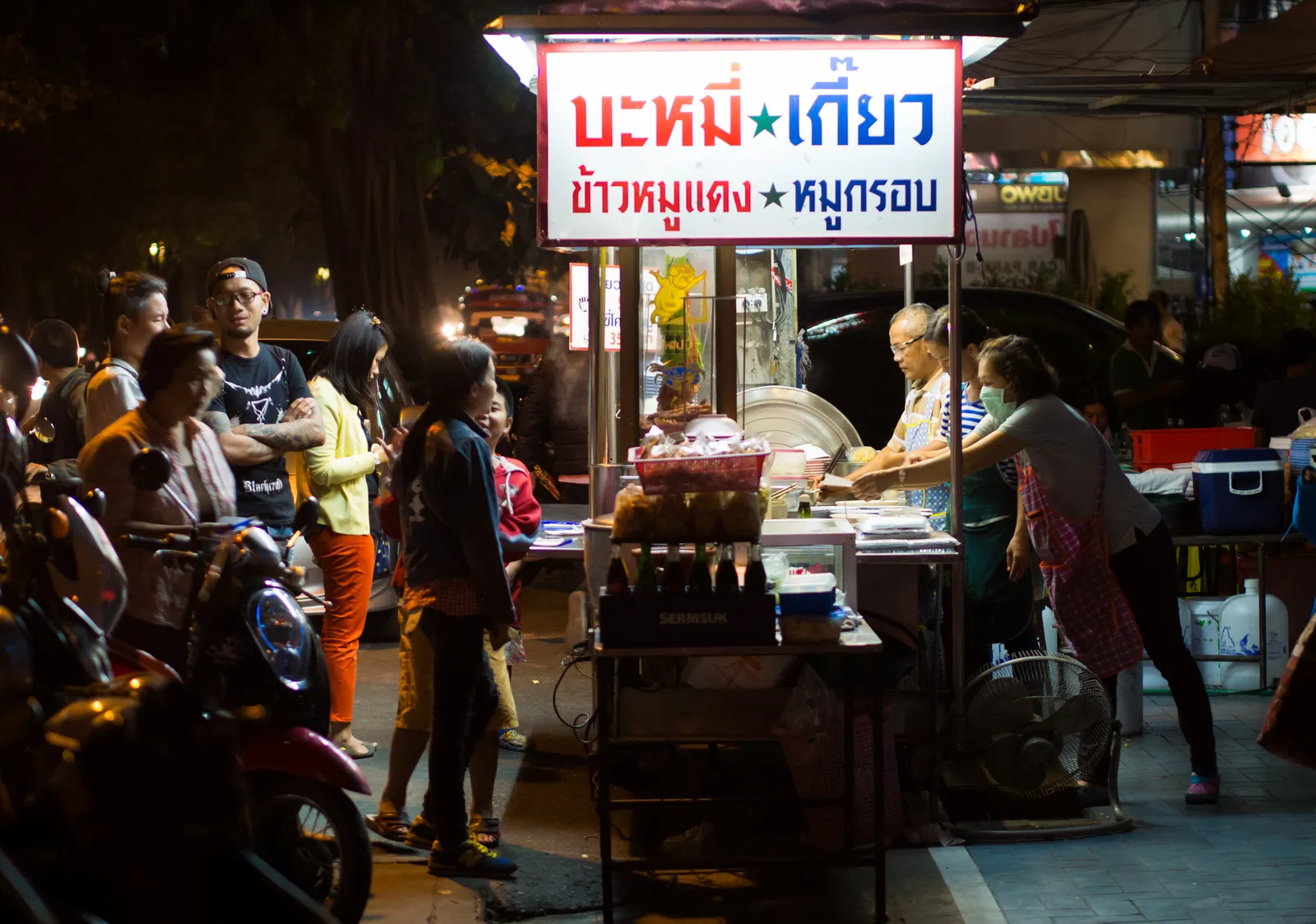 Night street food stall with a red Thai sign for noodles and dumplings, customers standing in front and a parked motorbike.