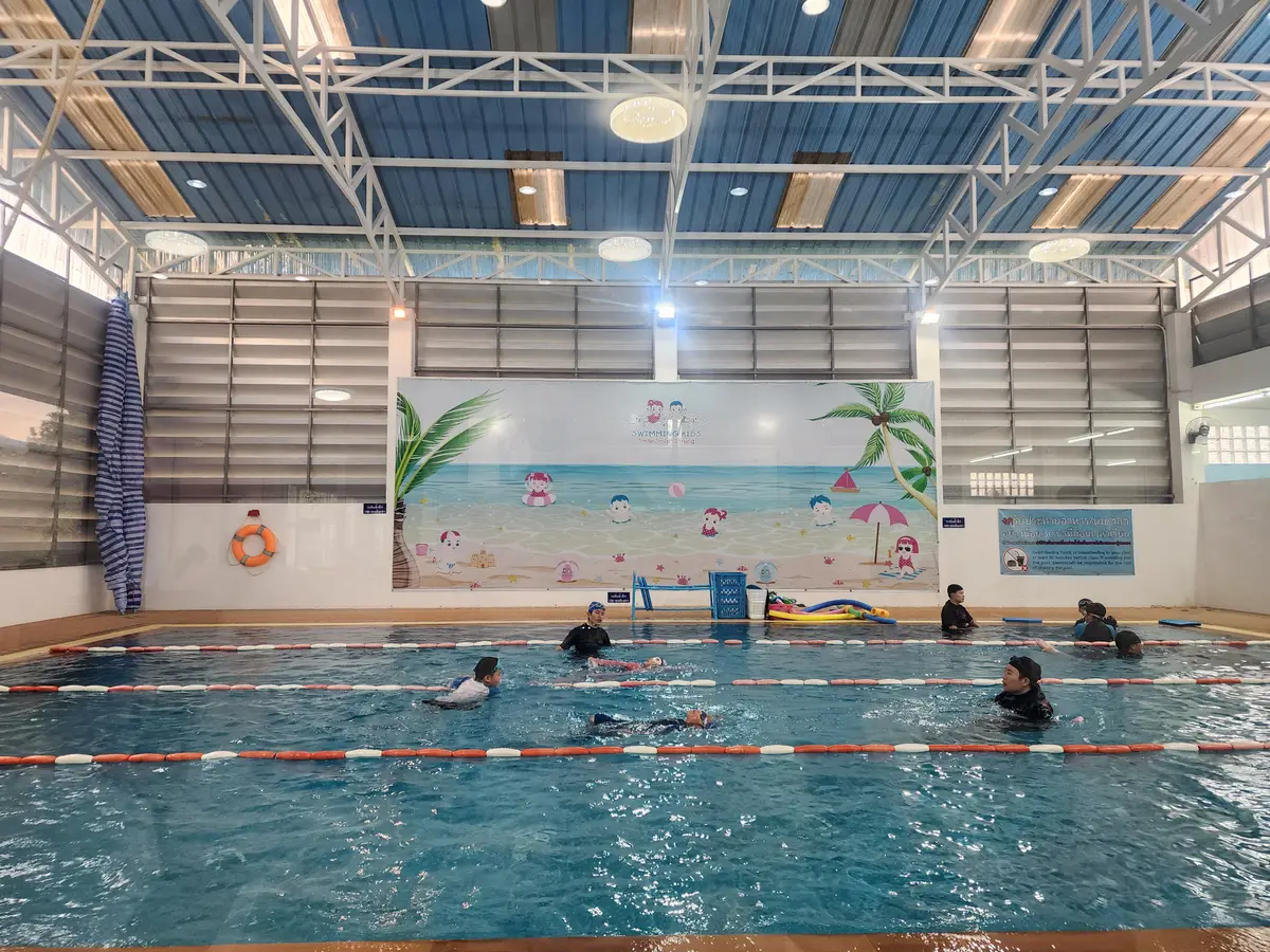 A covered indoor swimming pool with lane ropes, swimmers in the water and a beach mural on the far wall.