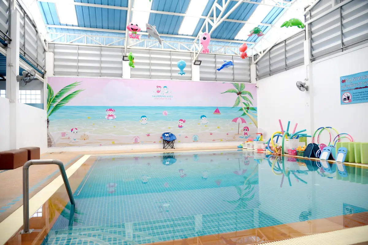 A shallow teaching pool in front of a pastel beach mural, with colourful pool noodles stacked at the side.