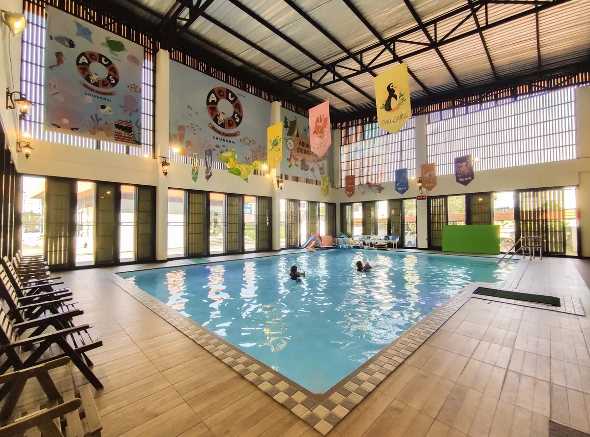 A tiled indoor pool under a high roof with sun loungers along one side and school banners on the wall.