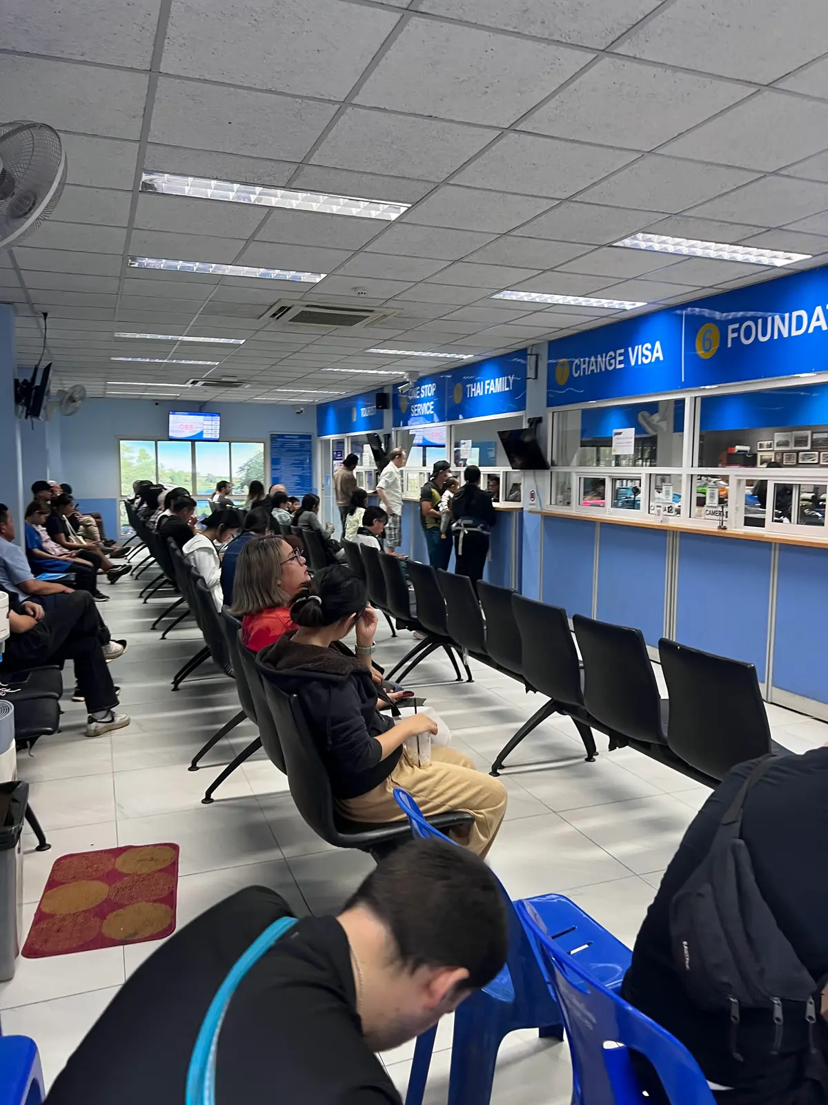 Visitor hall at Chiang Mai Immigration with blue service counters, a Change Visa sign and people waiting in chairs.