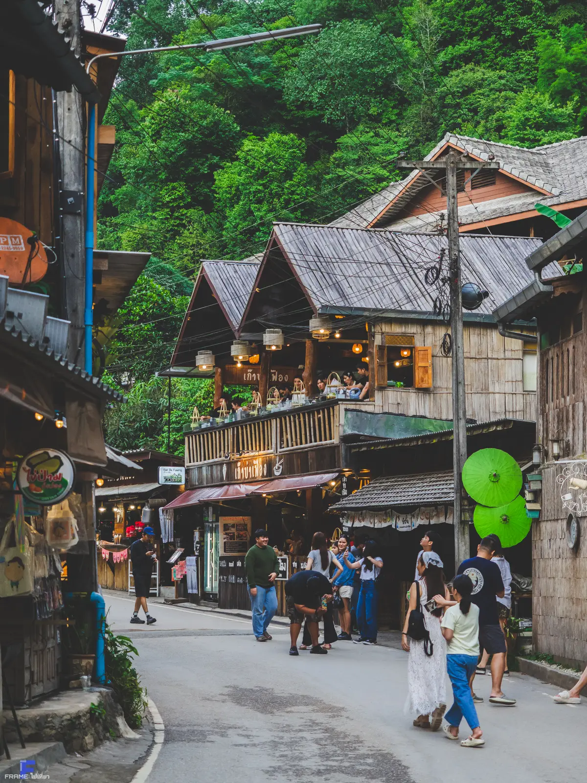 Village lane lined with wooden shops and cafes, with visitors walking along it and trees behind