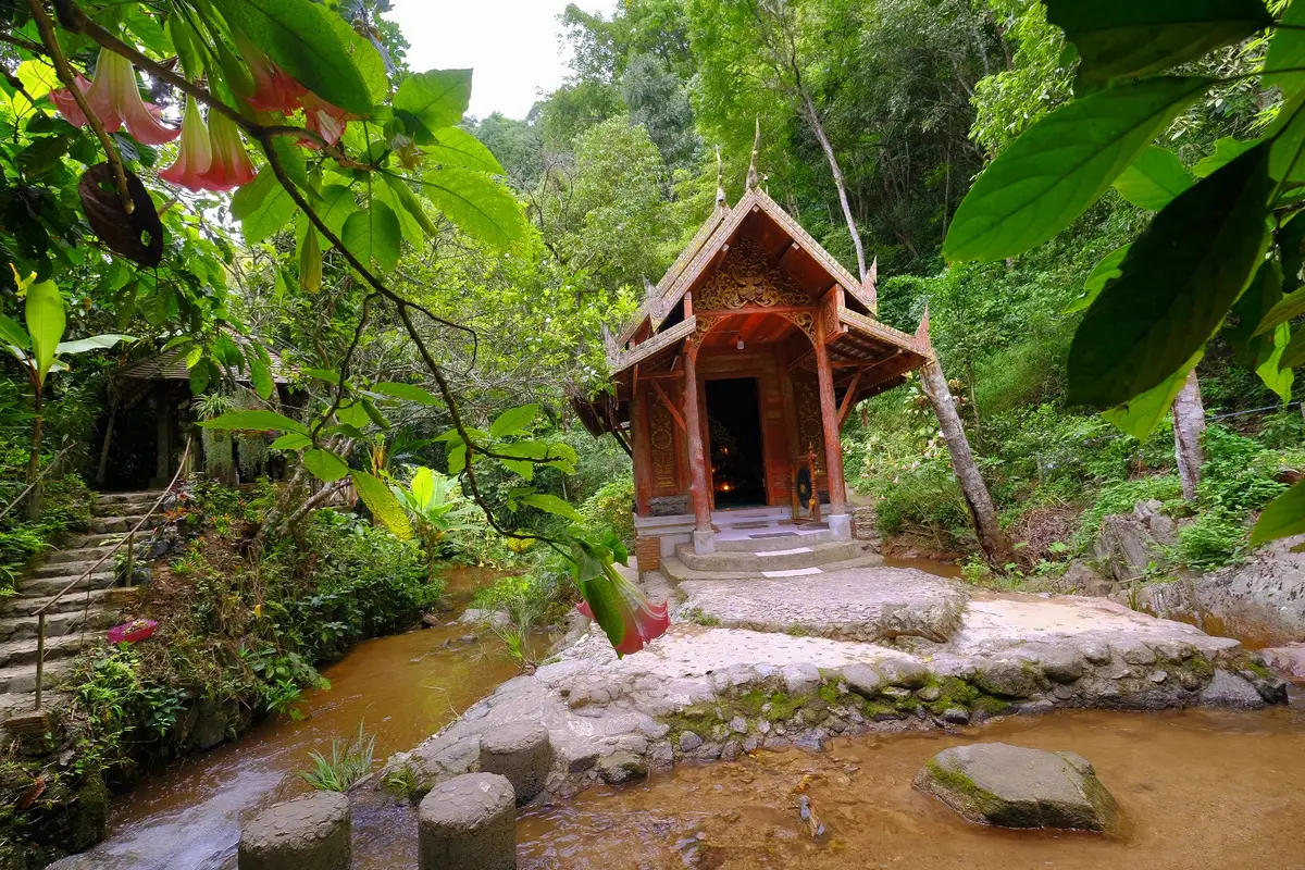 Small red wooden shrine pavilion beside a shallow stream among green leaves