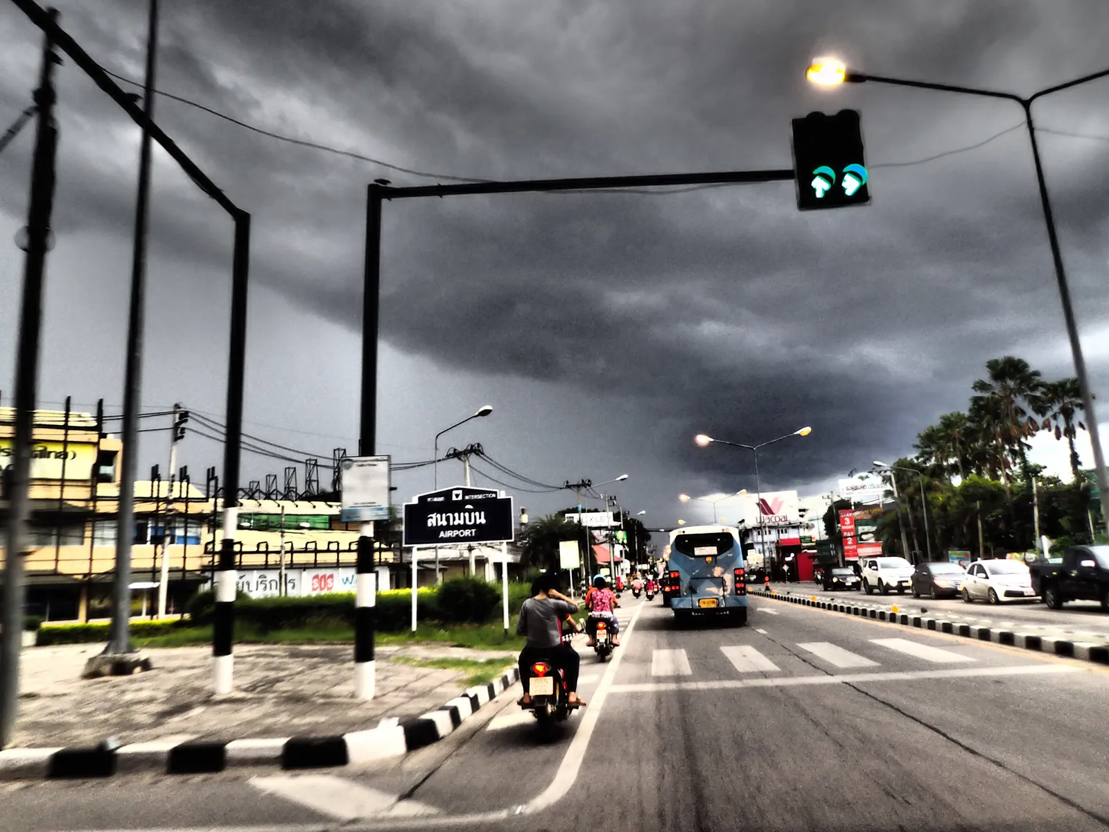 Road under a dark storm sky with a rider on a scooter in the left lane and a green traffic light ahead.