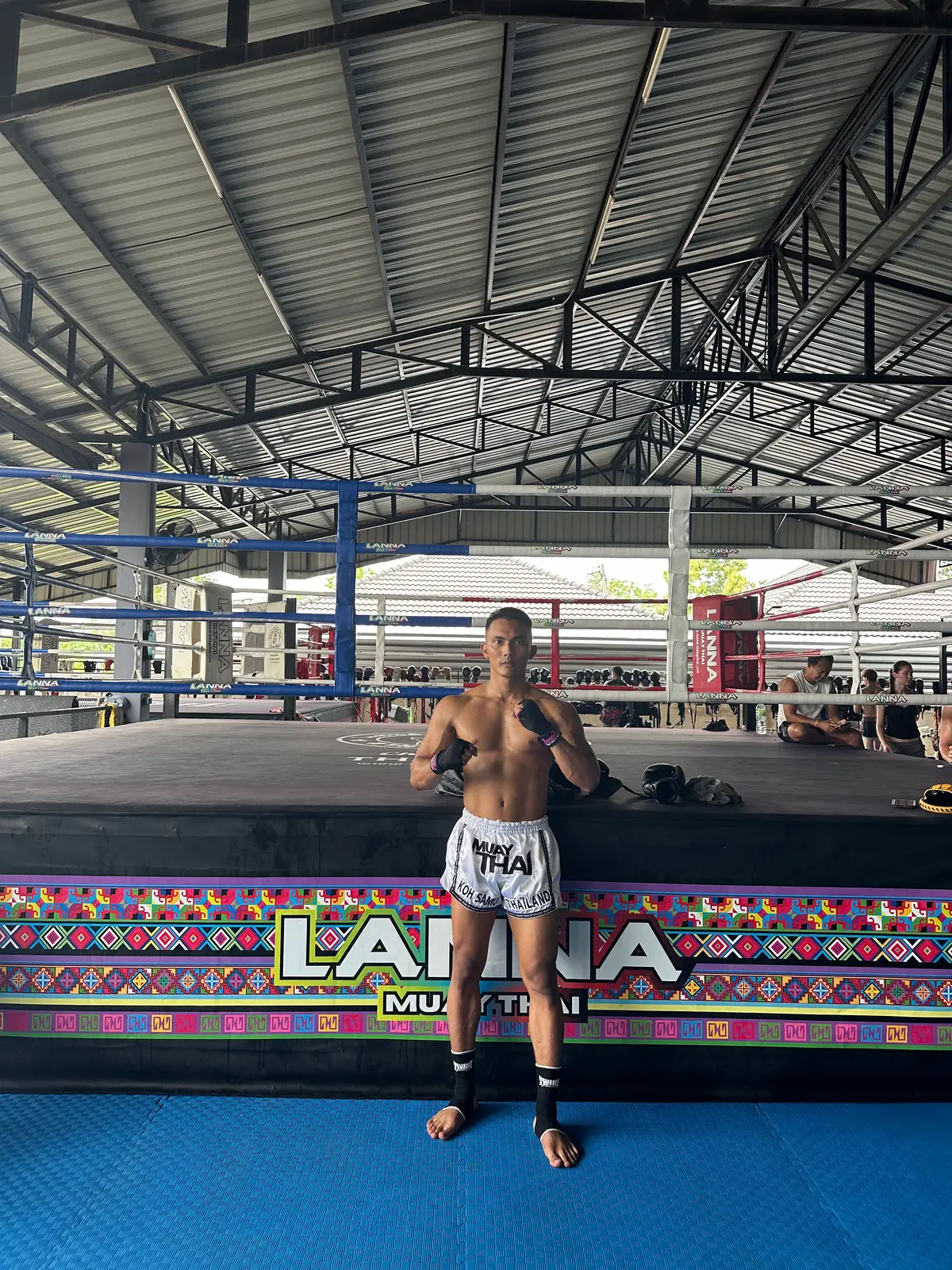 A fighter in shorts stands in front of a ring with a decorative skirt in a large steel-roofed hall.