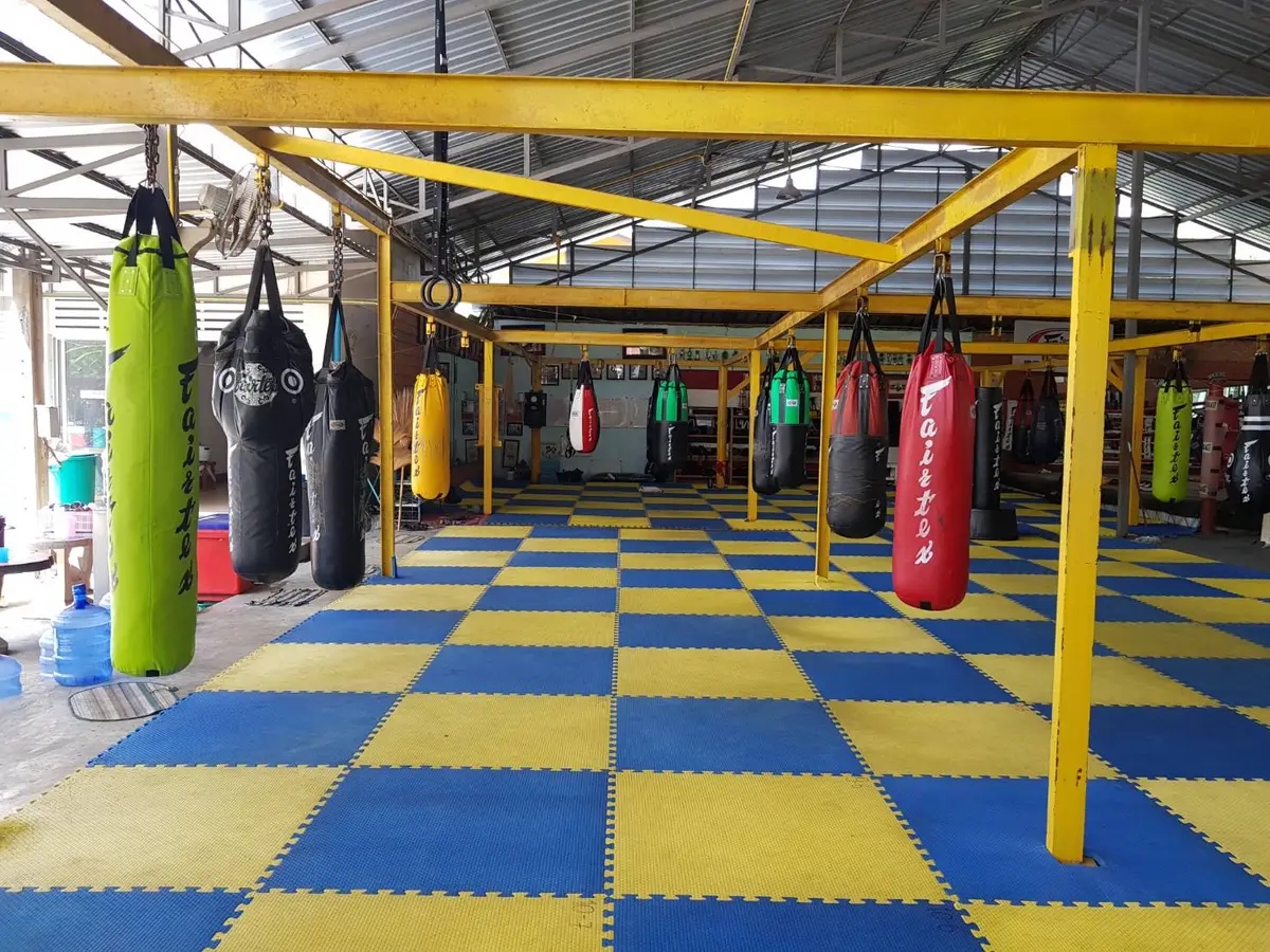 Covered gym floor with blue and yellow mats and rows of heavy bags hanging from yellow frames.
