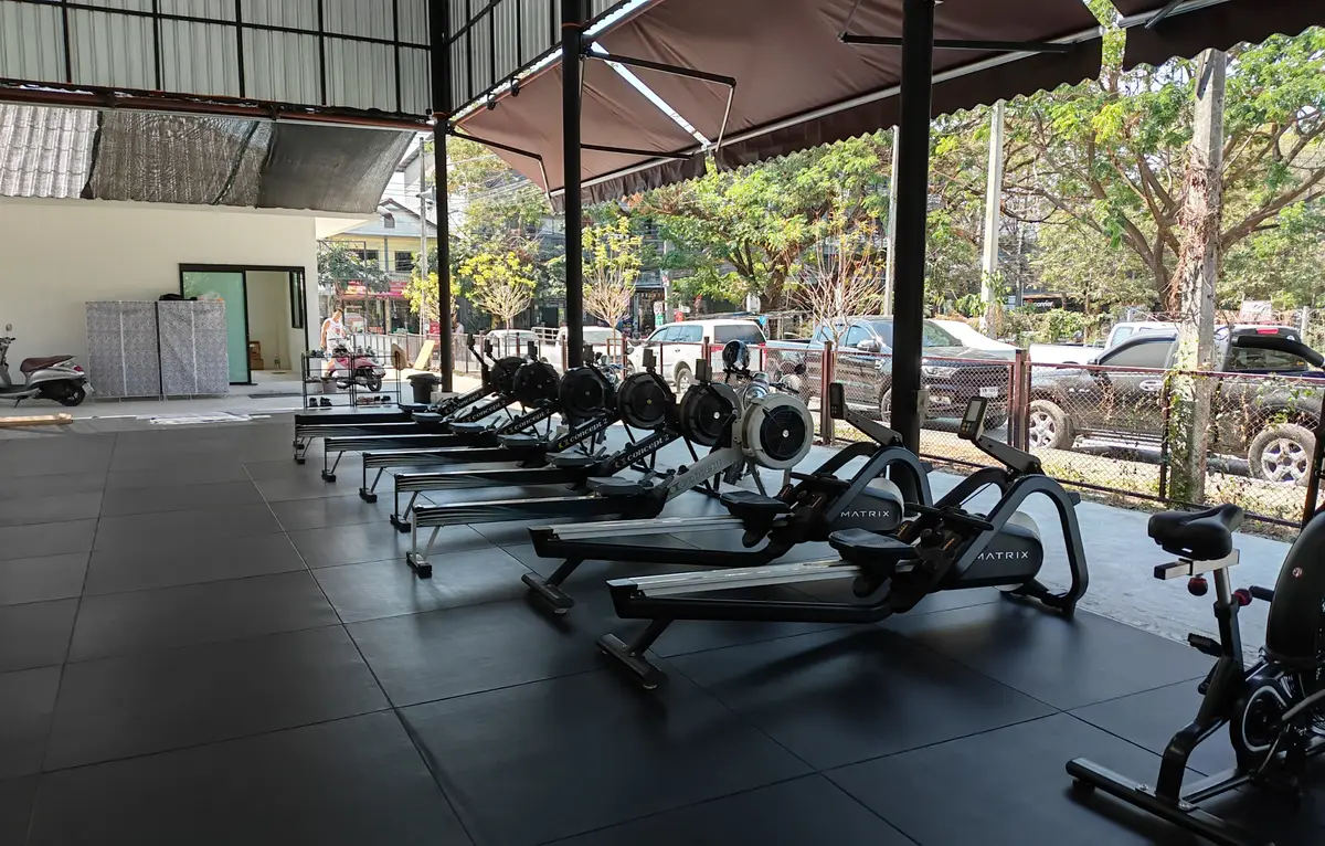 Row of rowing machines on a rubber floor under a steel roof, with cars parked outside.
