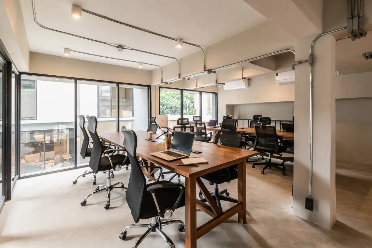 Long wooden desk with office chairs and laptops beside tall windows