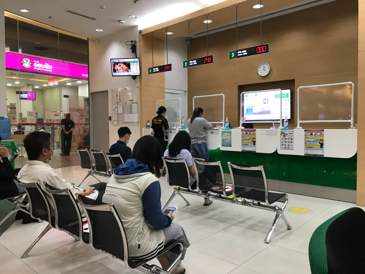Customers seated on benches in a bank hall with queue number displays above the counters.