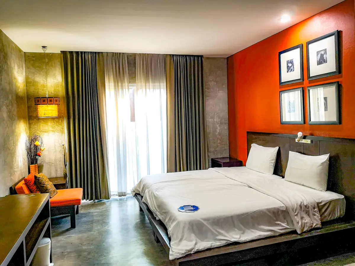 Hotel room with a double bed, an orange accent wall with framed pictures and long curtains