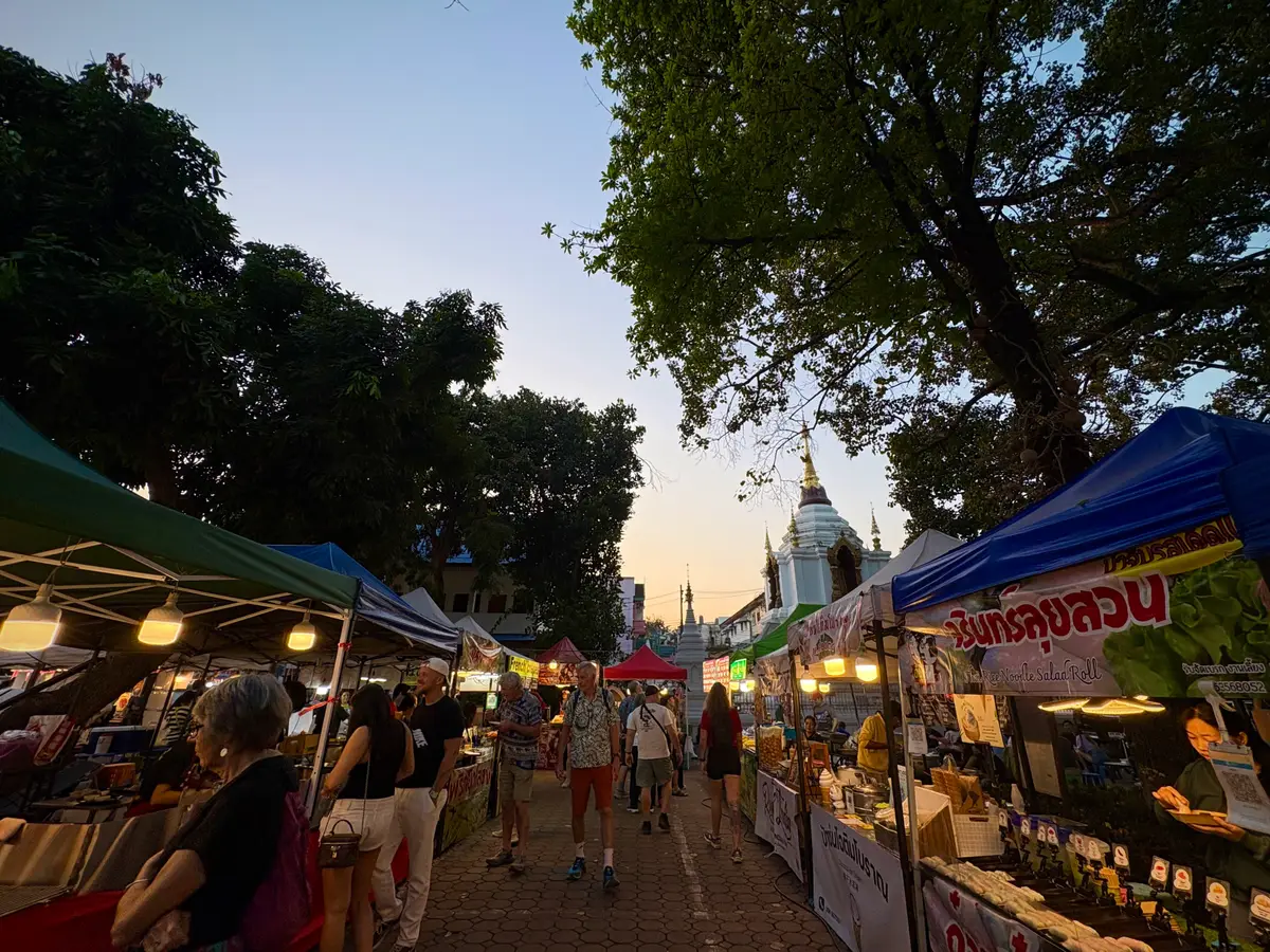 Rows of market tents along a tree-lined road at dusk, with a temple roof in the distance.