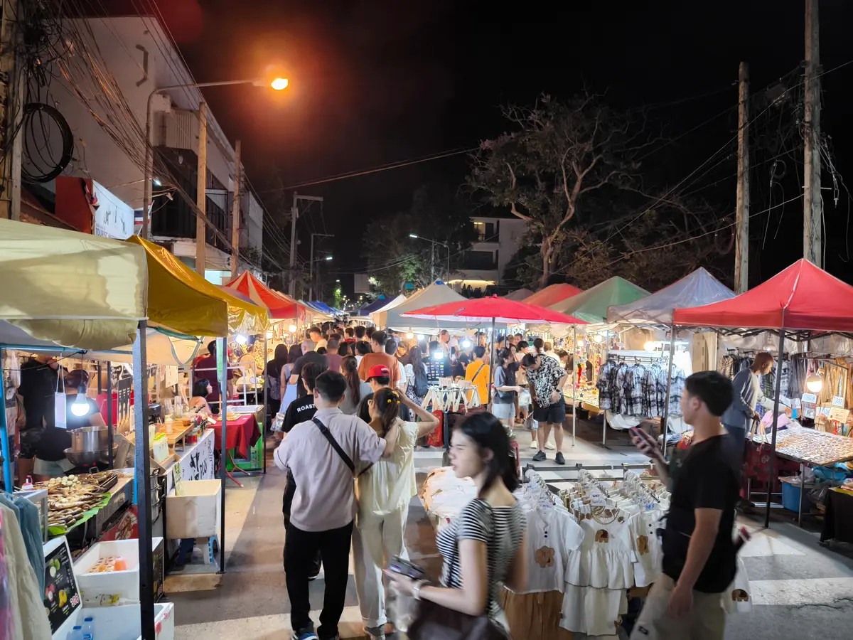 Brightly lit night market stalls with clothes and food and a crowd browsing between them.