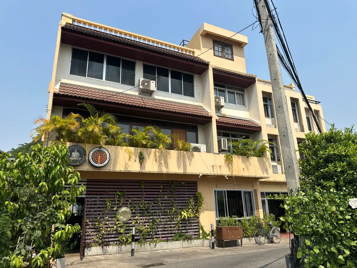 A pale multi-storey building with balconies and potted plants beside trees.