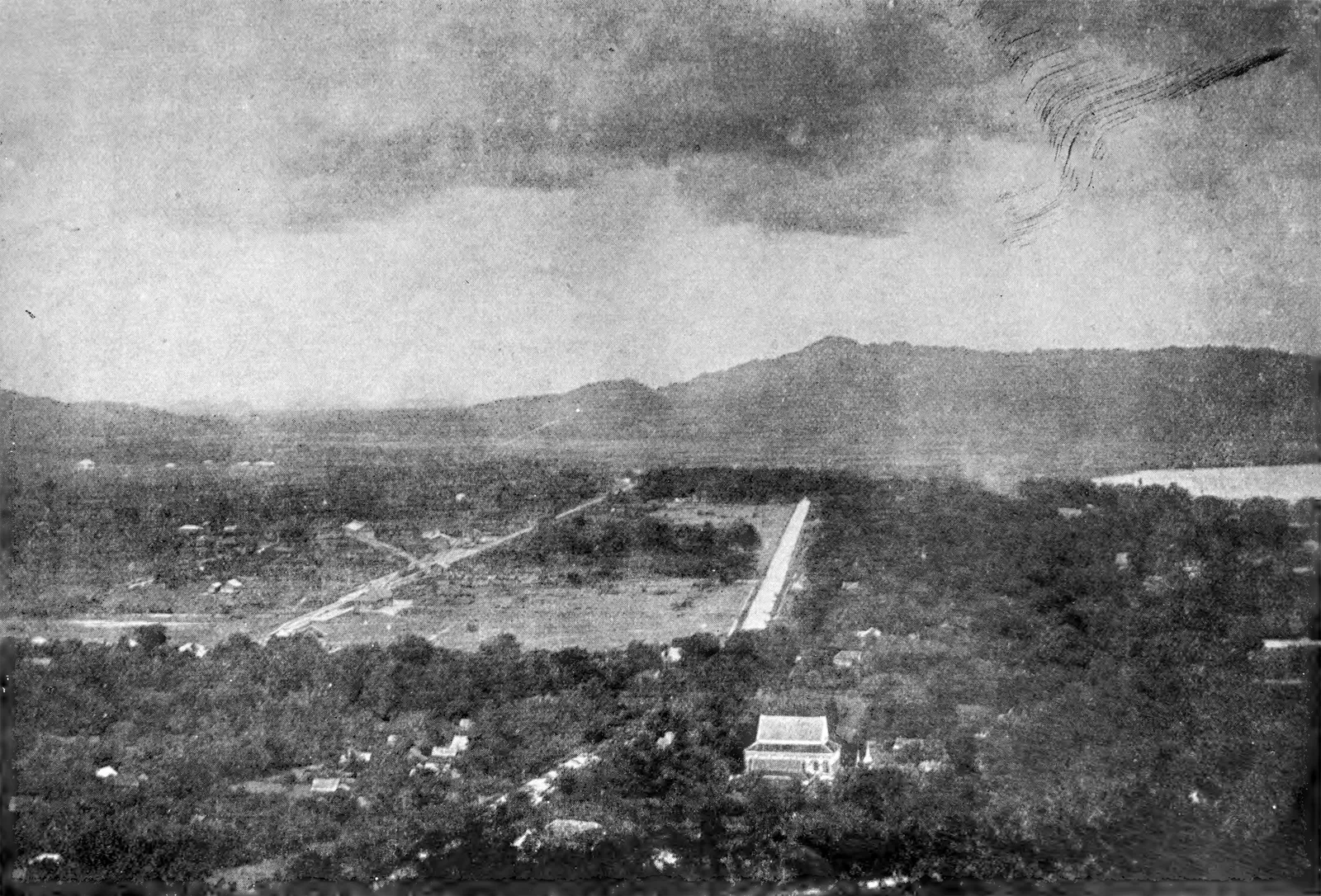 Old black-and-white photograph of a wooded plain with a long straight road and a mountain range under cloud.