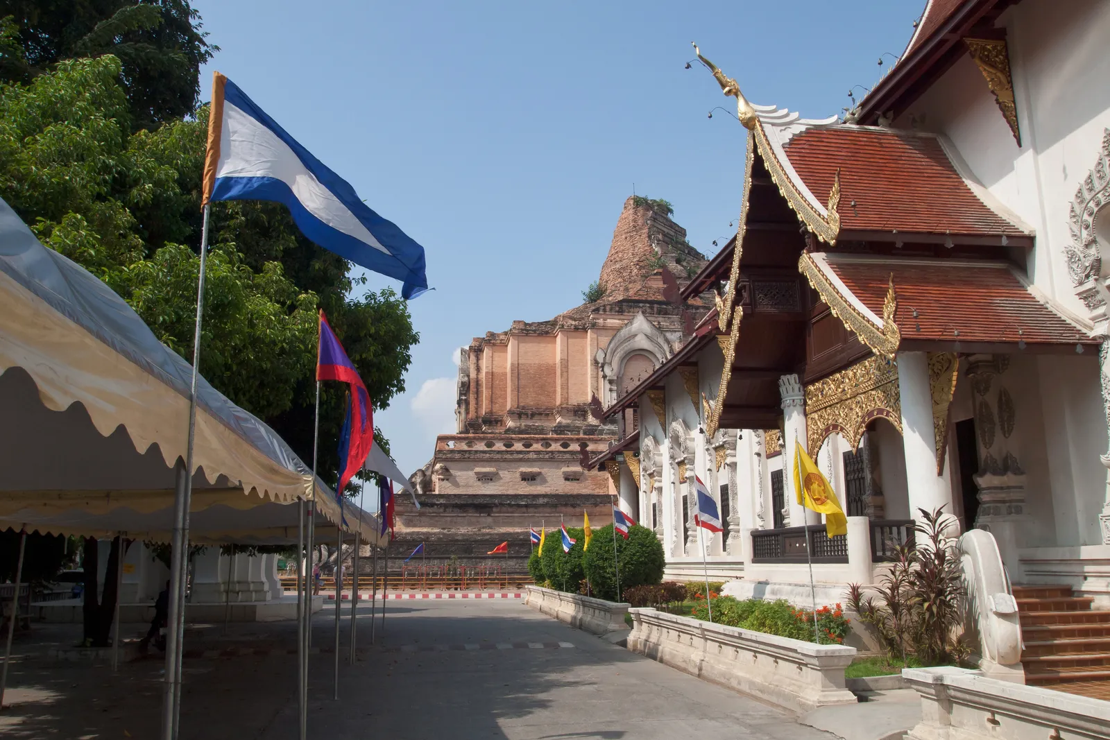 Path through the temple grounds with flags, a white hall on the right and the brick chedi in the distance