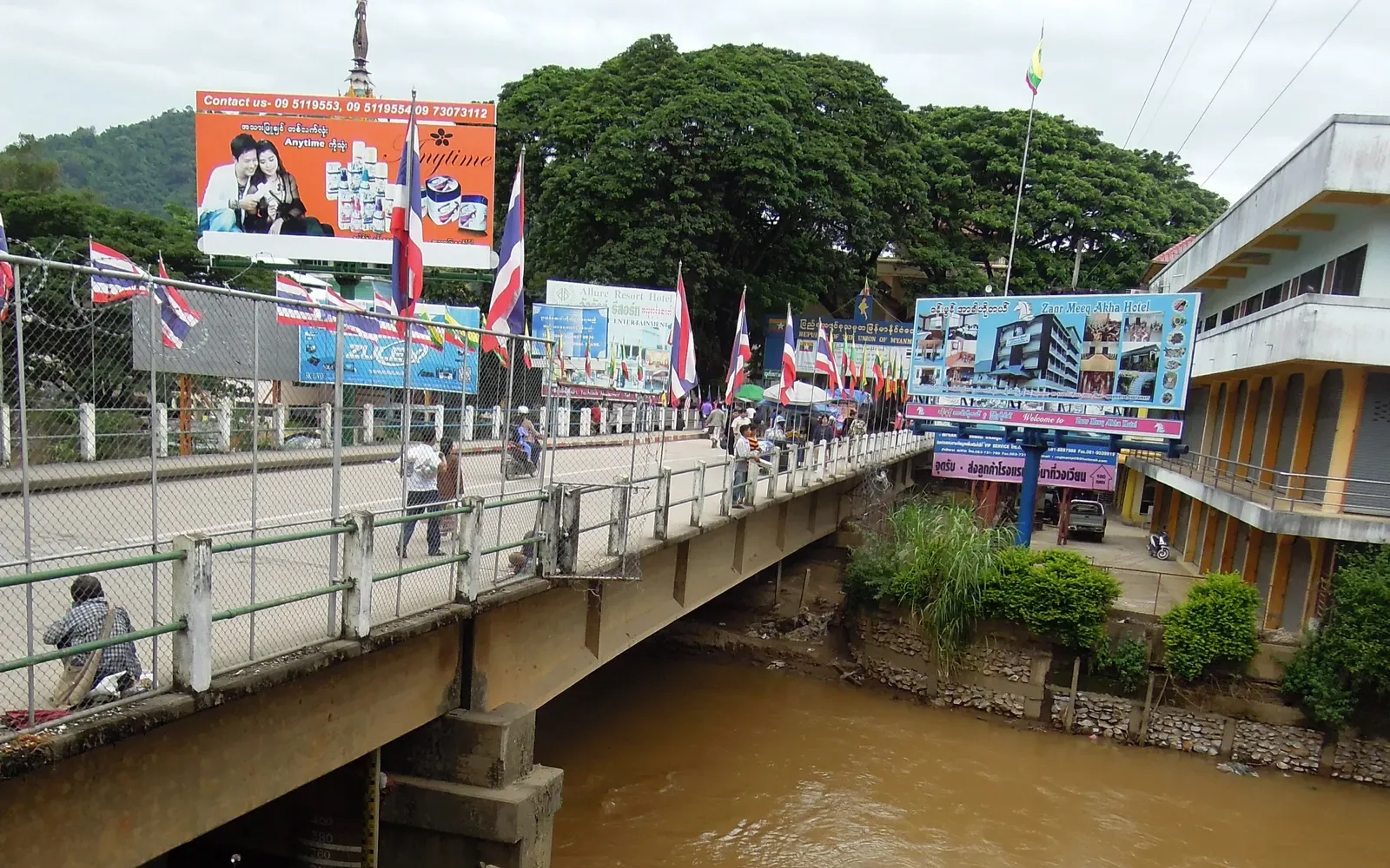 Road bridge over a brown river, with flags, billboards and a parked car beside it.