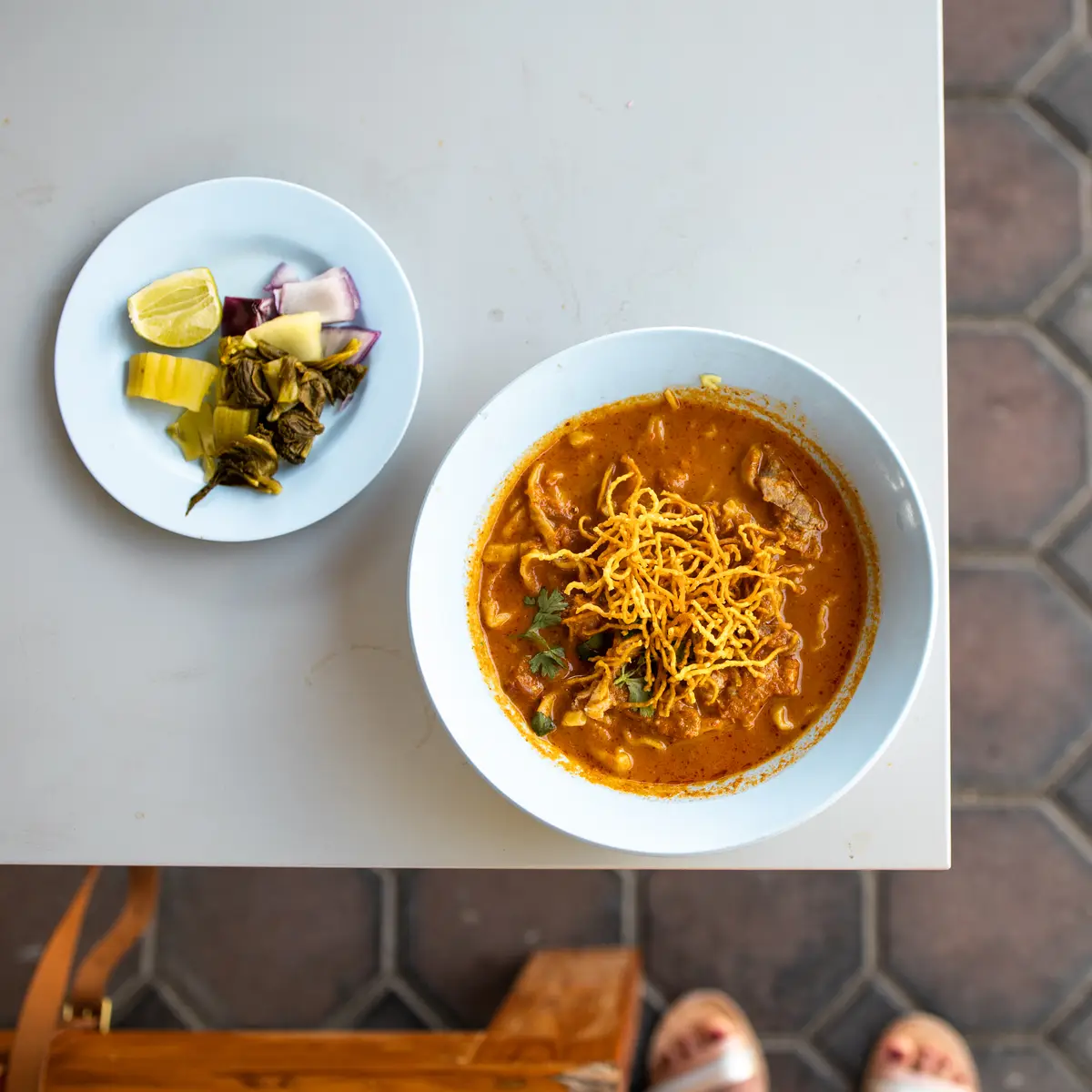 A bowl of khao soi with crispy noodles on top, with a side plate of pickled greens, shallots and lime.