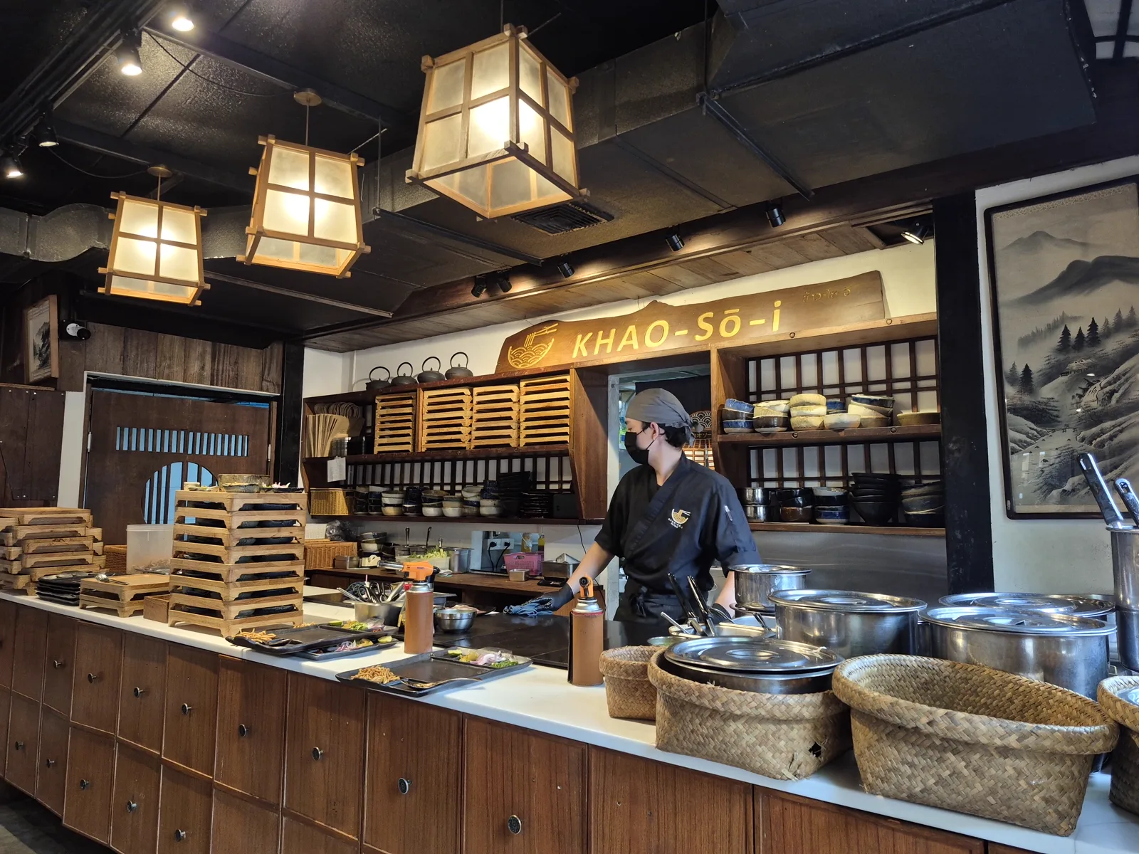Open kitchen counter with a Khao-So-i sign, a staff member in black, hanging lanterns and steel pots.